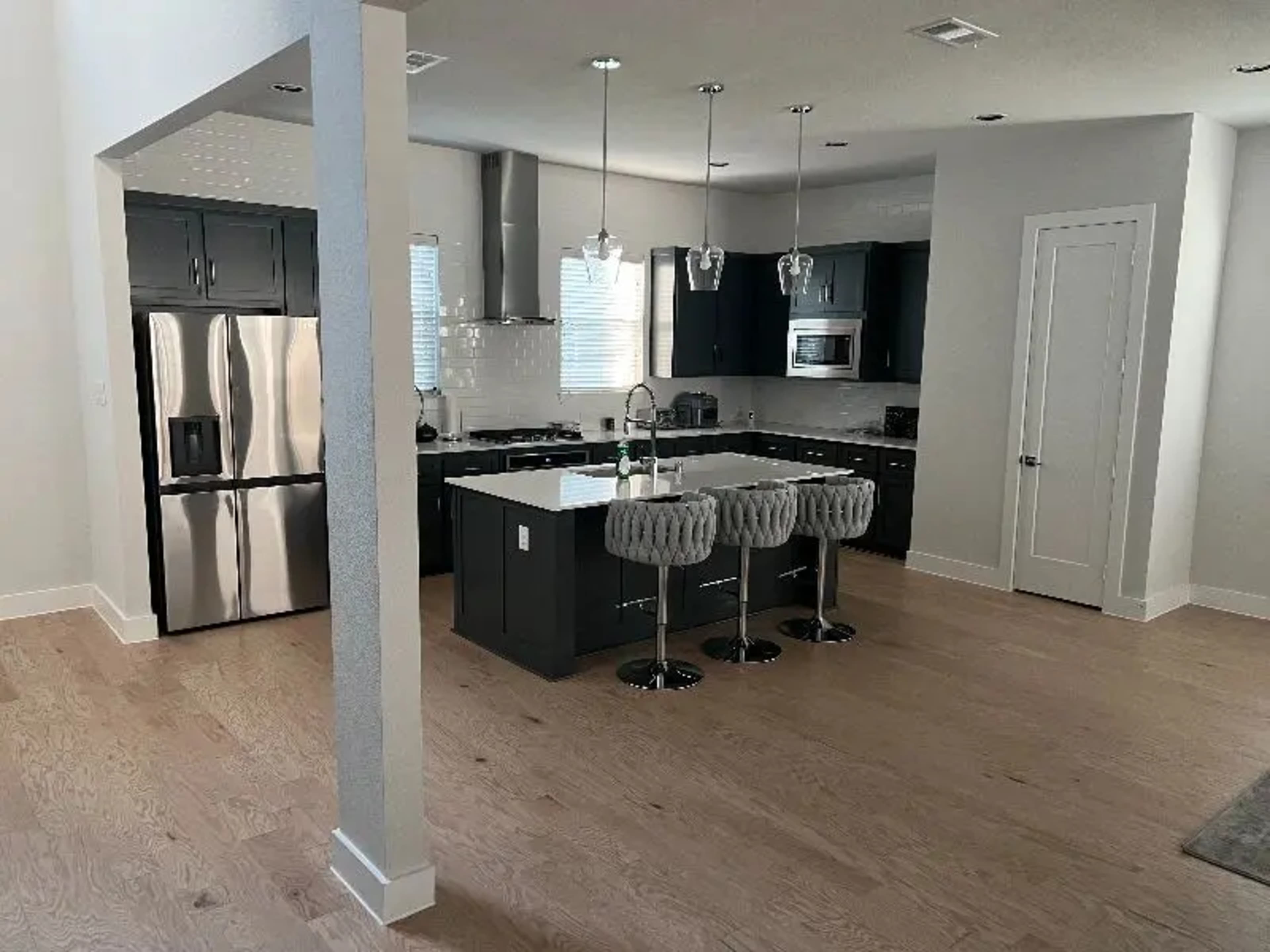 A modern kitchen features dark cabinets, stainless steel appliances, a central island with three bar stools, and light hardwood flooring.
