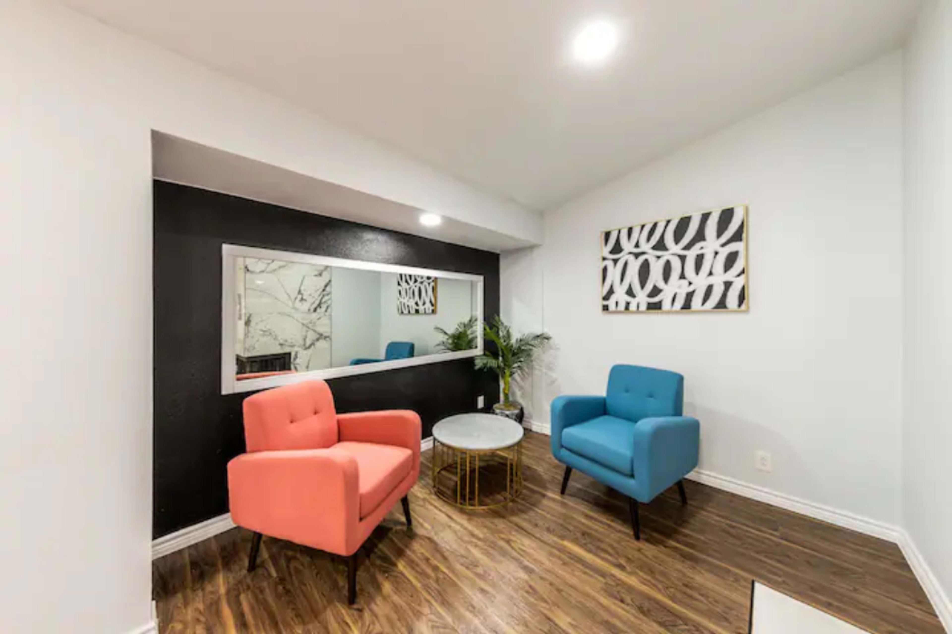 A small room featuring two colored chairs, a round table, a large mirror, and a piece of wall art.
