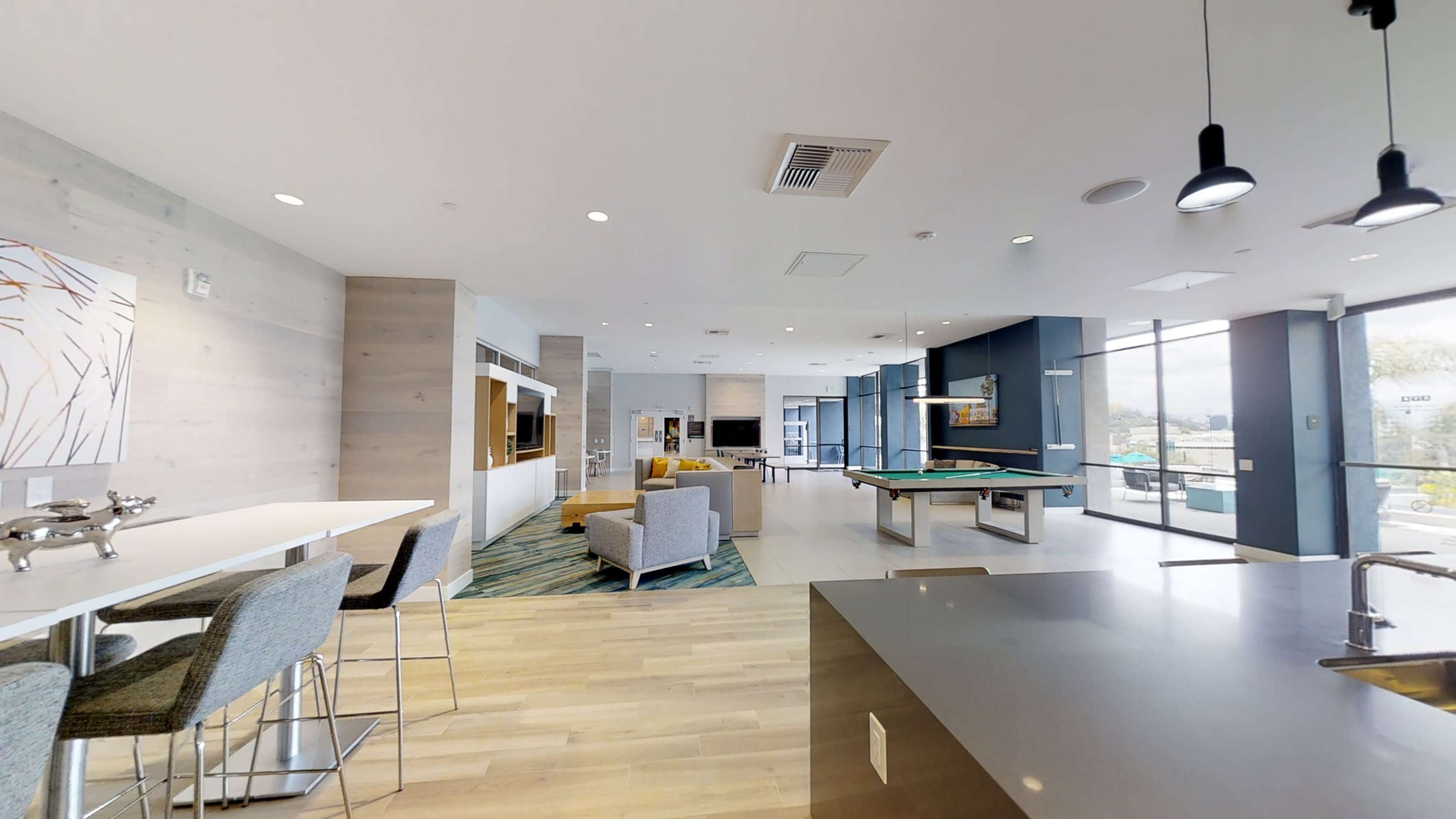 The image shows a modern recreational room with seating areas, a pool table, and large windows providing natural light.