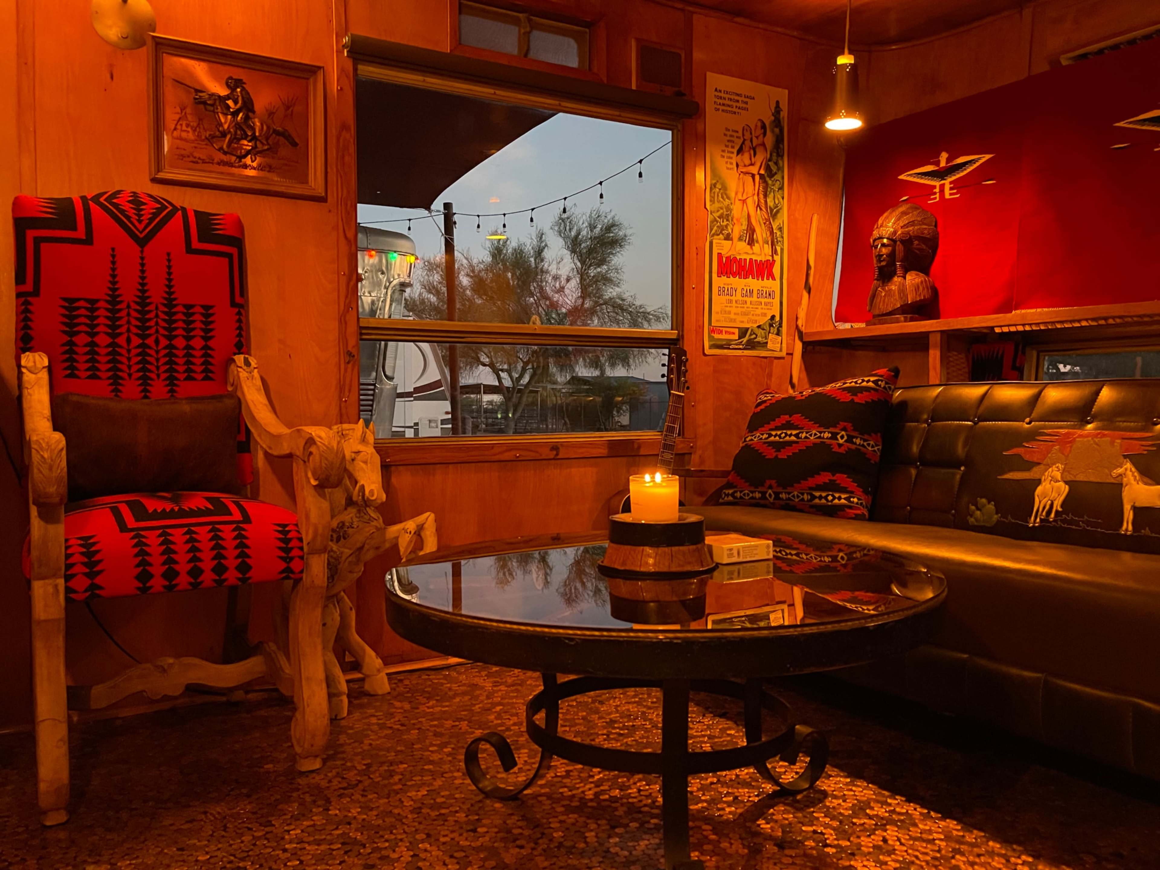 The image shows a cozy interior space with wooden walls, a circular coffee table, a black leather couch adorned with colorful cushions, and artistic decor, including posters and a large chair.