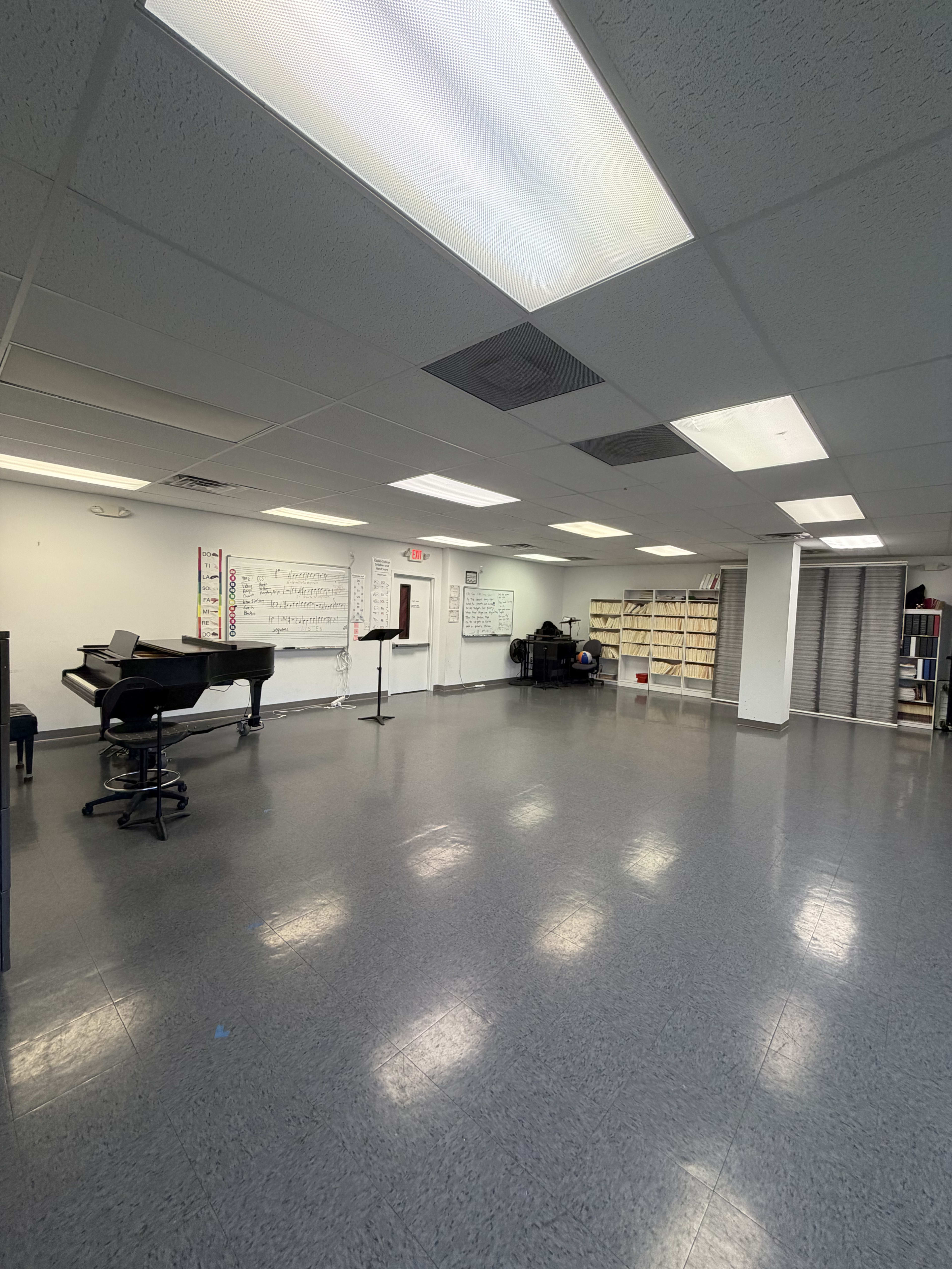 The image shows a spacious music room with a grand piano, a music stand, whiteboards, and shelves filled with books in a well-lit setting.
