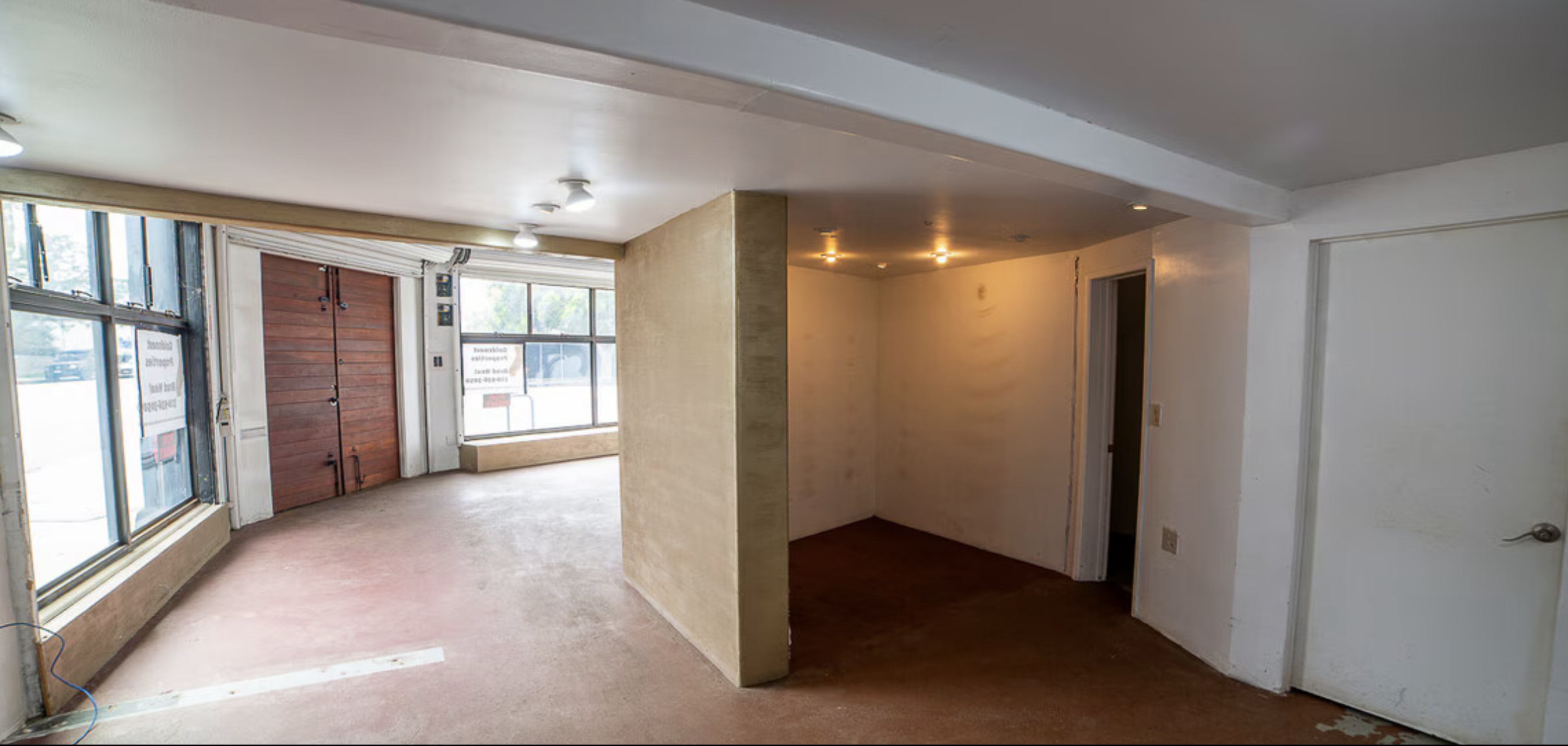 The image shows an empty room with bare walls, a concrete floor, and large windows along one side.