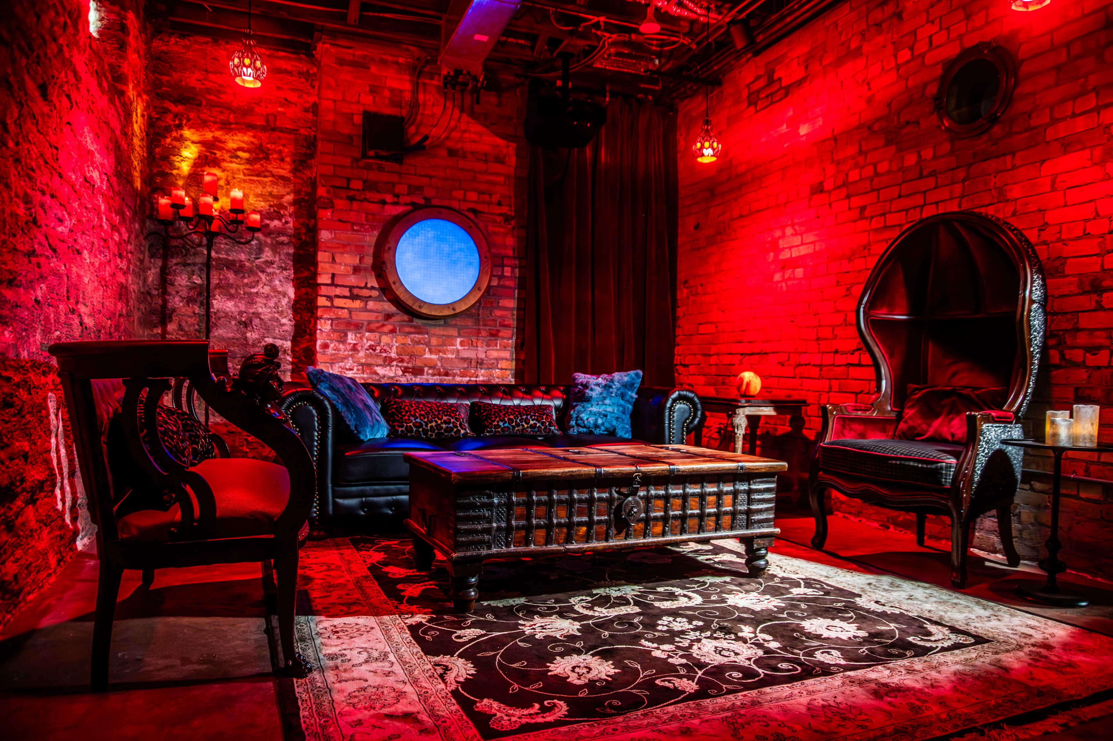The image depicts a dimly lit lounge with exposed brick walls, featuring ornate furniture, a large round mirror, and a decorative rug at the center.