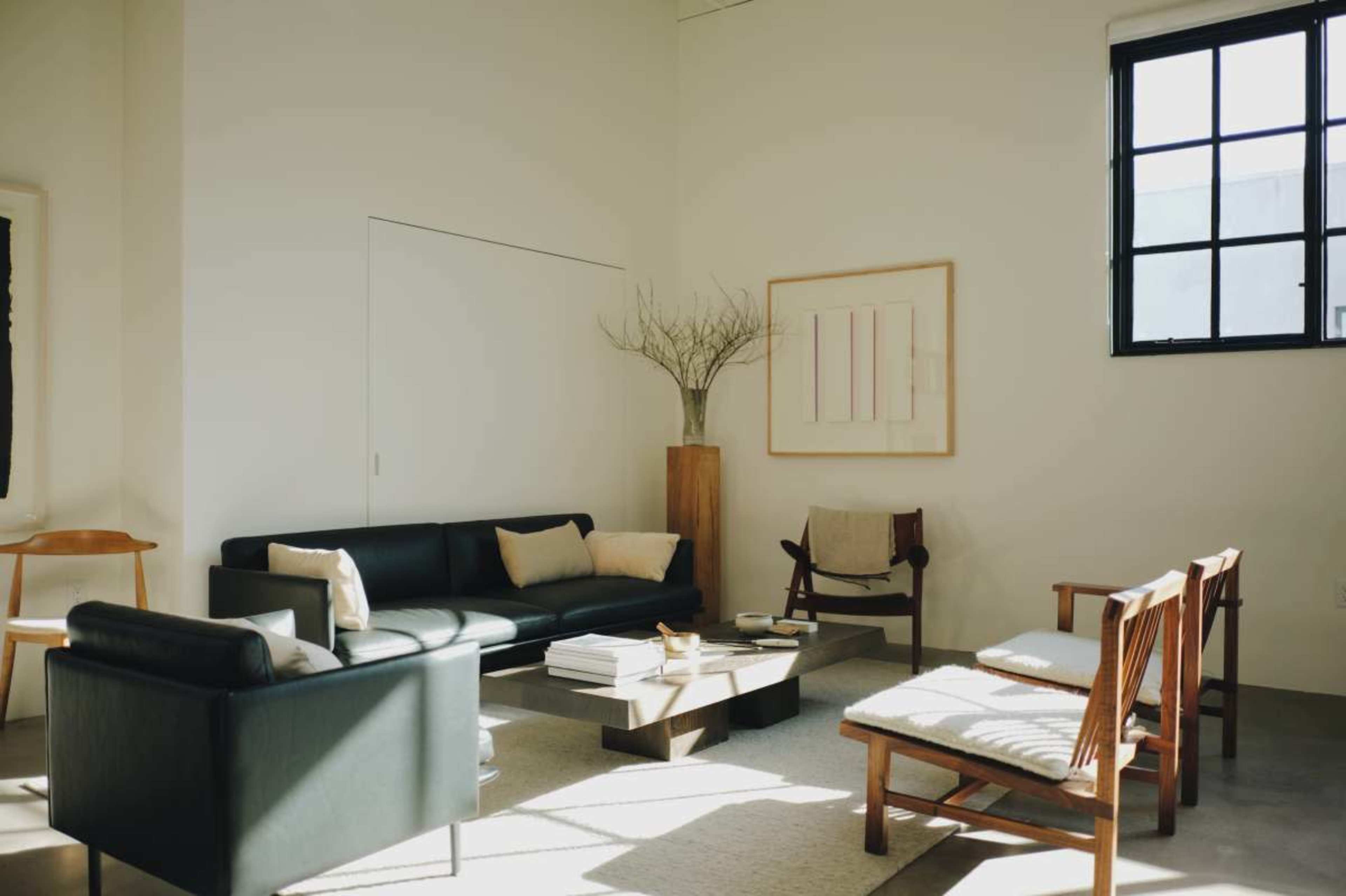 The image shows a minimalist living room featuring a black sofa, wooden chairs, and a coffee table, with natural light coming in through a large window.