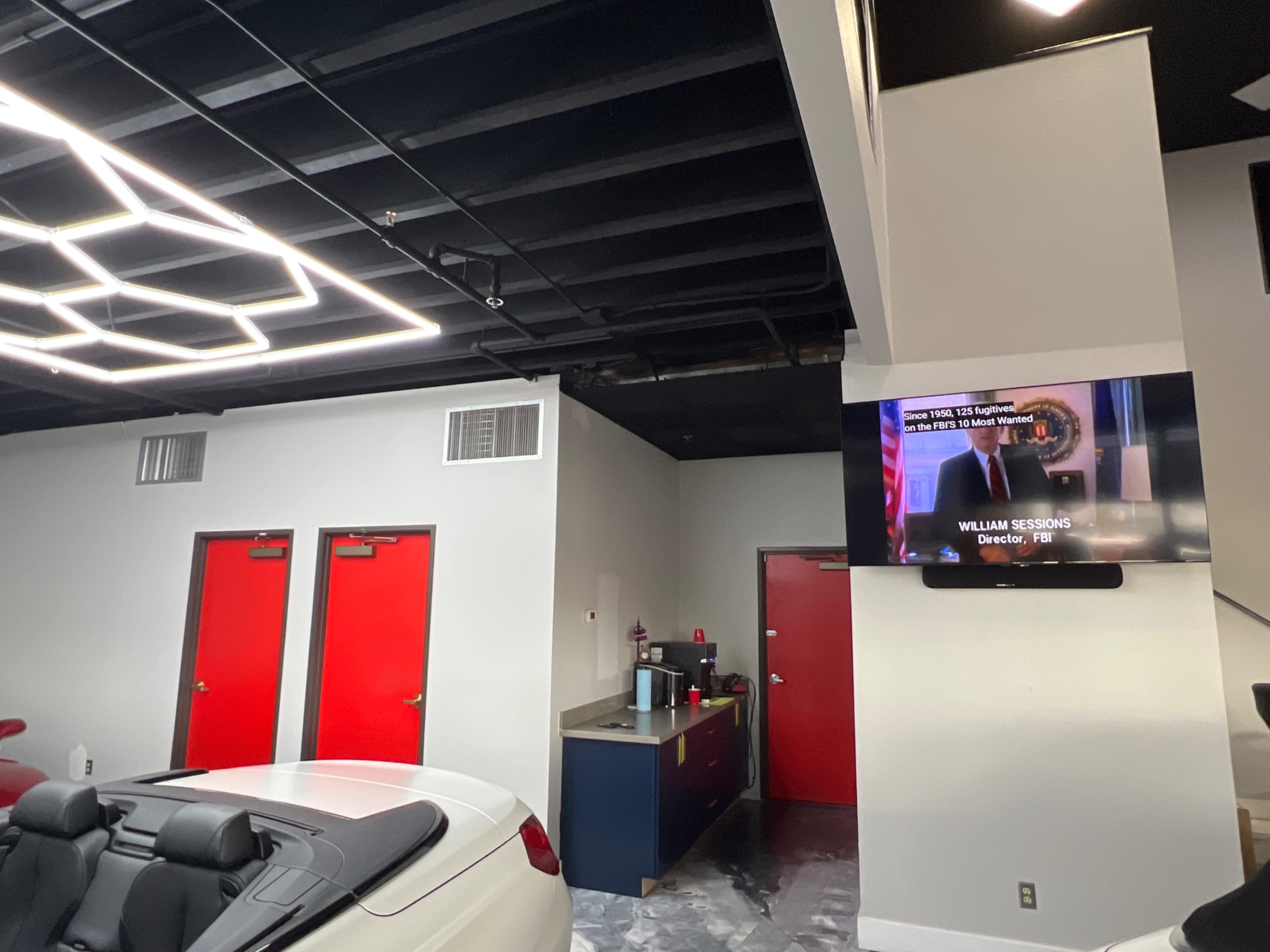 The image shows an interior space with a white convertible car in the foreground, red doors on the left, and a television mounted on the wall displaying a news segment.
