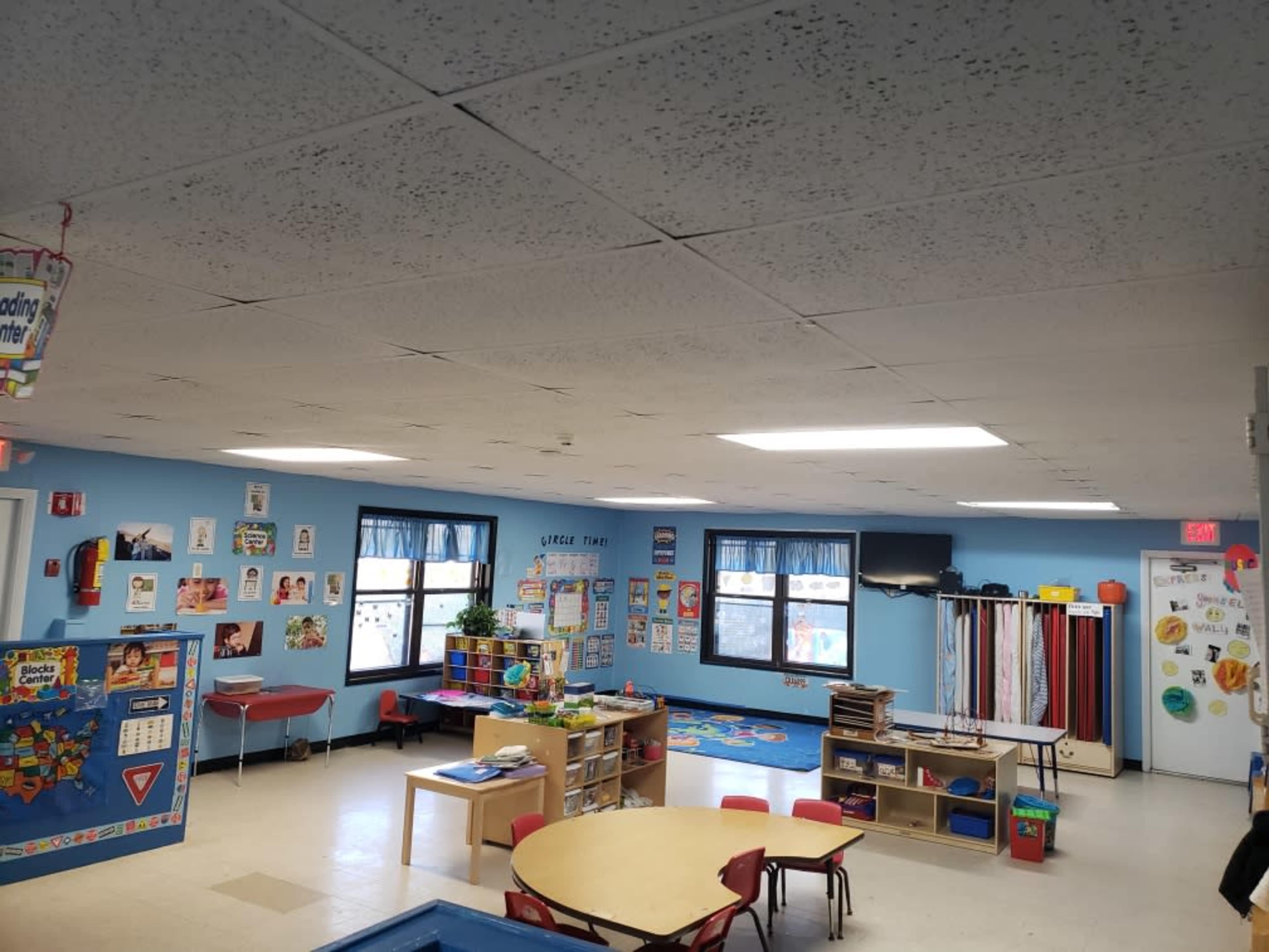 A brightly lit classroom features colorful educational posters, tables, chairs, and various learning materials scattered throughout the space.