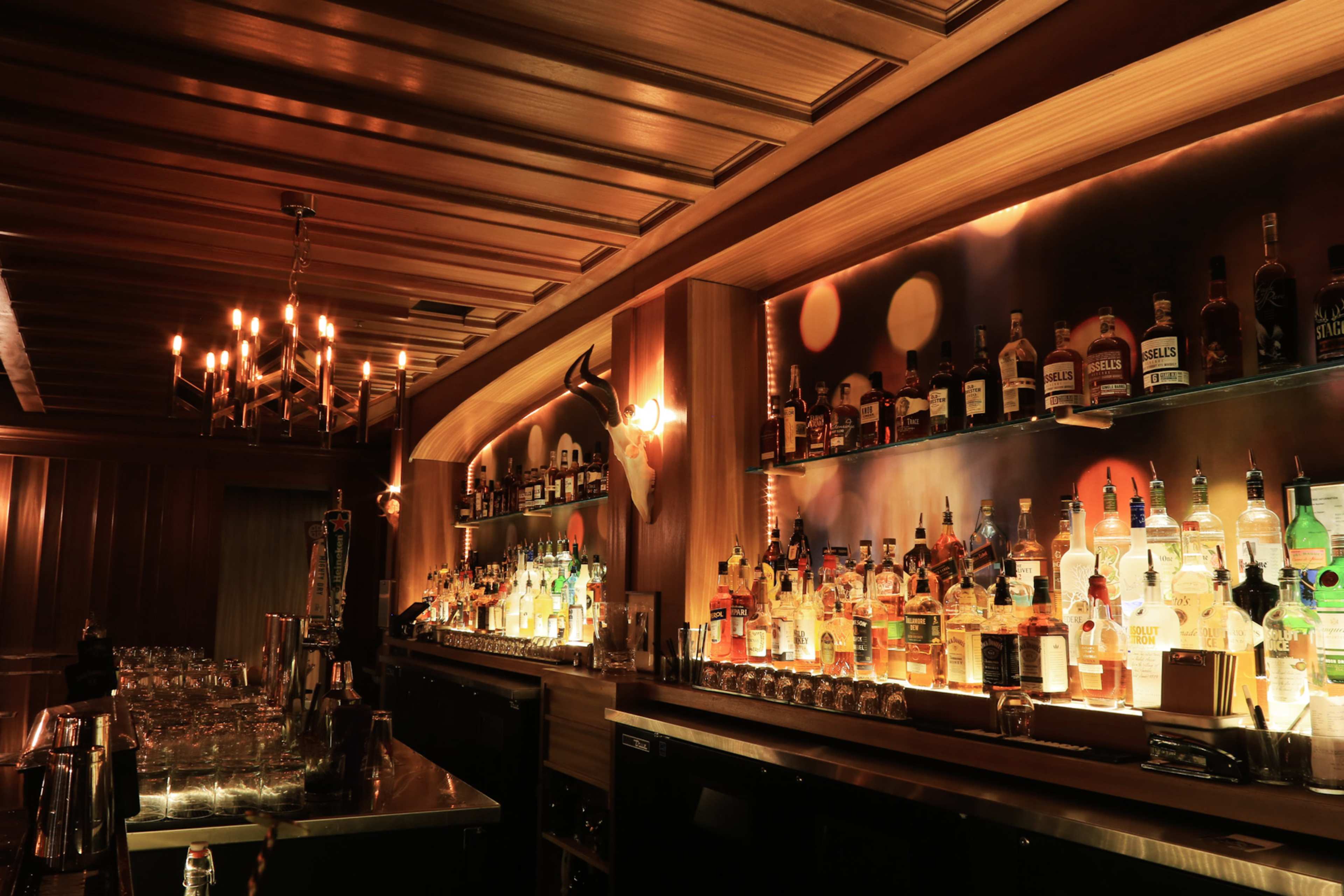 A dimly lit bar with shelves lined with various bottles of liquor, a chandelier overhead, and a sleek countertop.