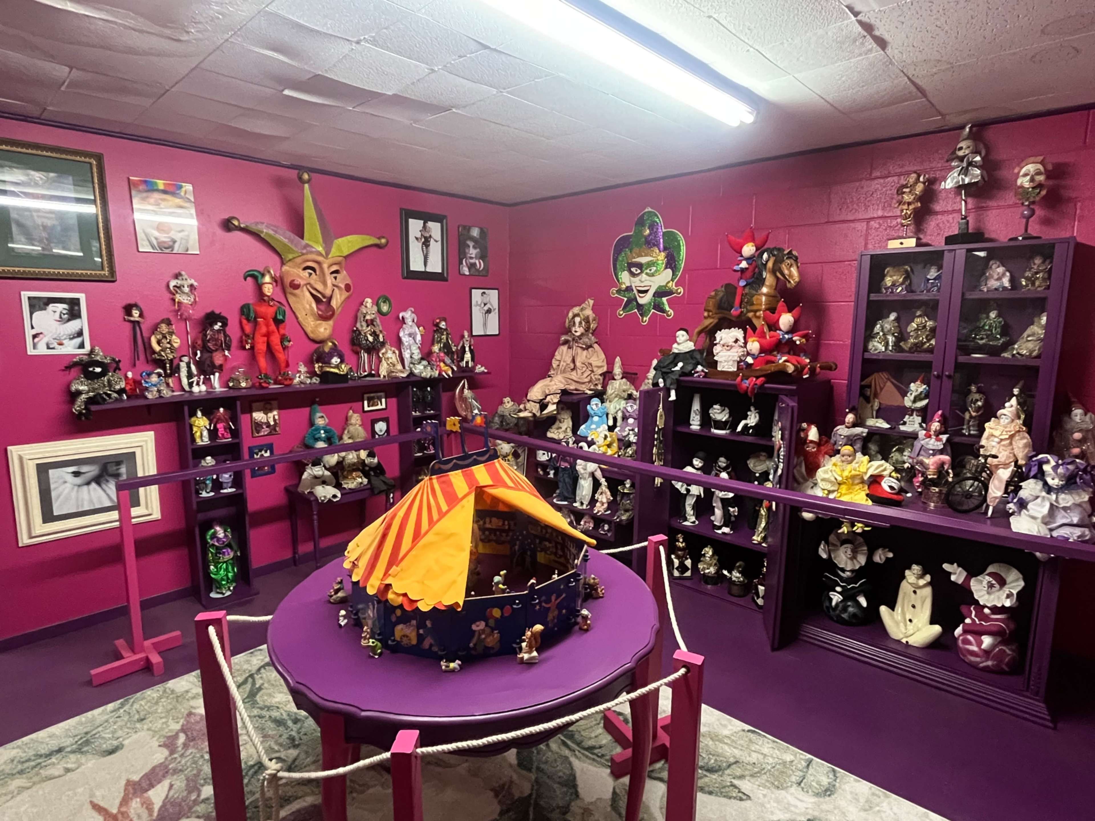 The image shows a vibrant room with pink walls filled with various colorful dolls, masks, and decorative items displayed on shelves and a circular table.