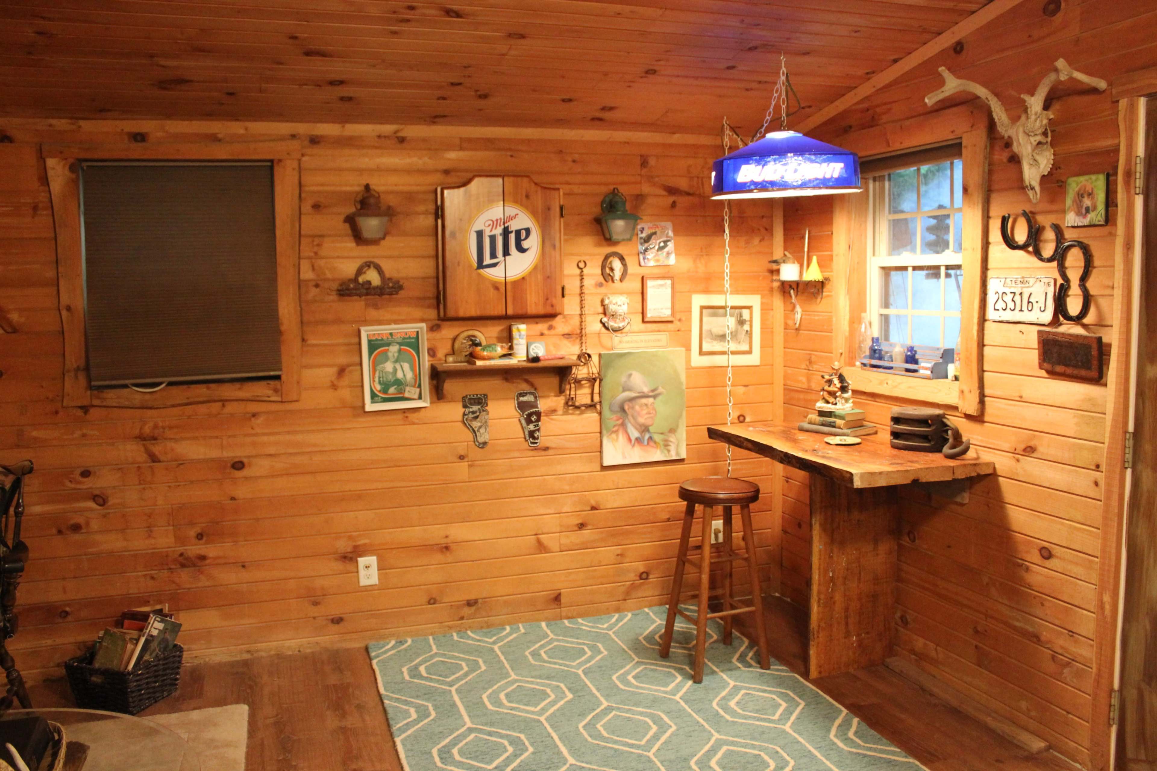 The image shows a wooden-paneled room featuring a small bar area, a blue bar light, framed pictures on the walls, and a patterned rug on the floor.