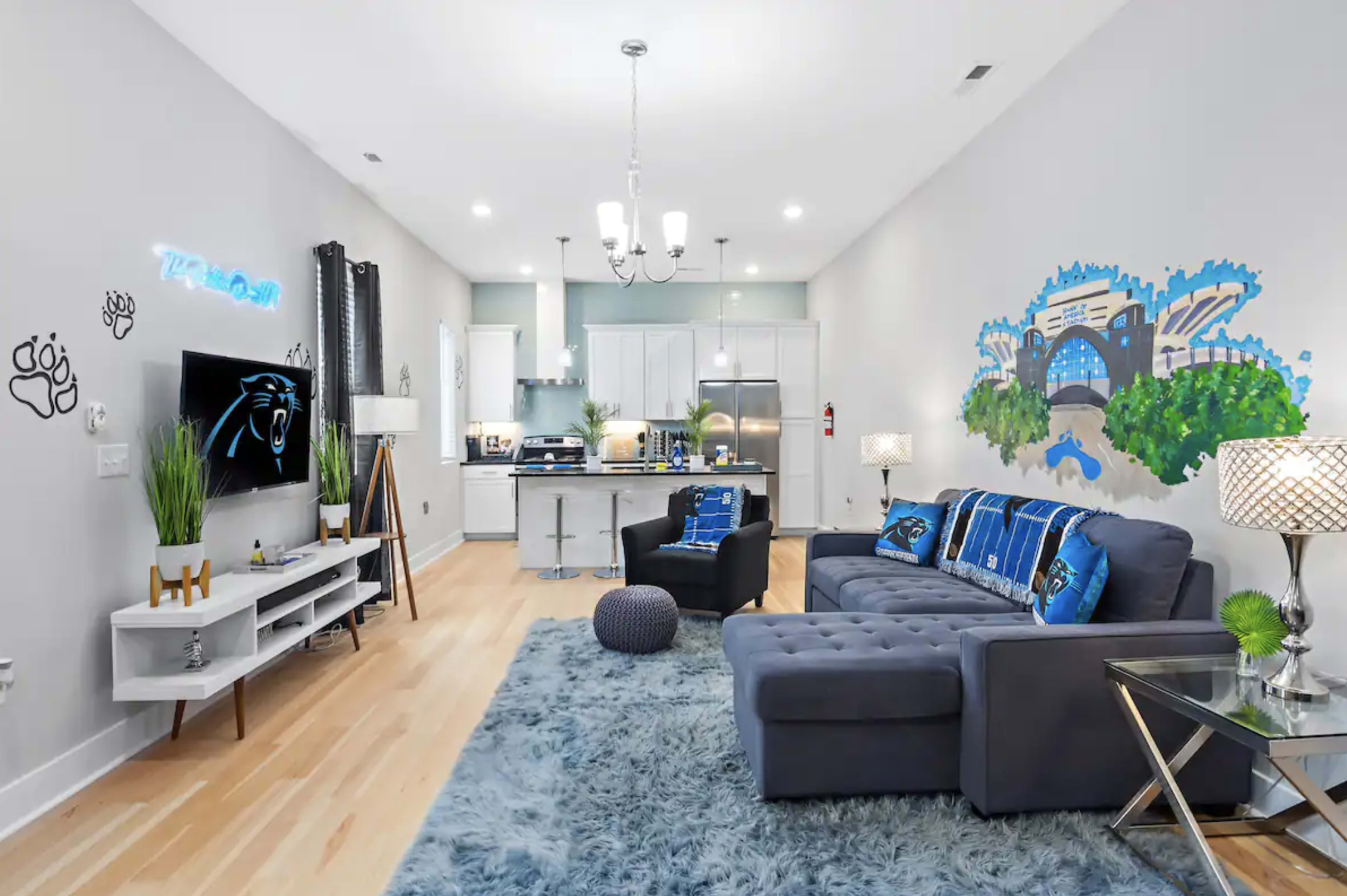 A modern living room with a blue-themed decor, featuring a sectional sofa, a large area rug, and a mural on the wall, all adjacent to a kitchen with white cabinetry.