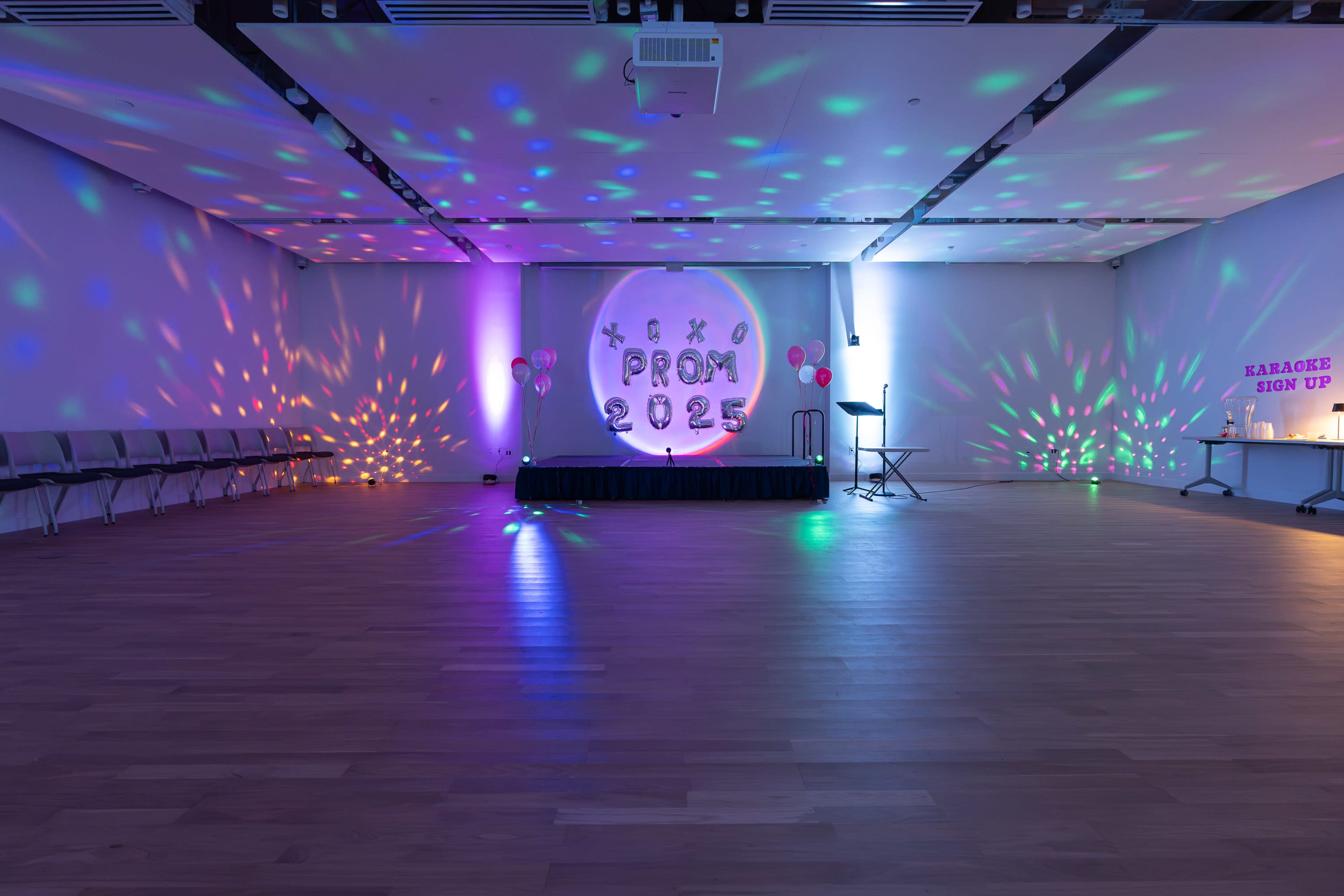The image shows a decorated event space for a prom, featuring a circular backdrop with "PROM 2023" written on it, colorful lights projected on the walls, and a stage with a microphone.