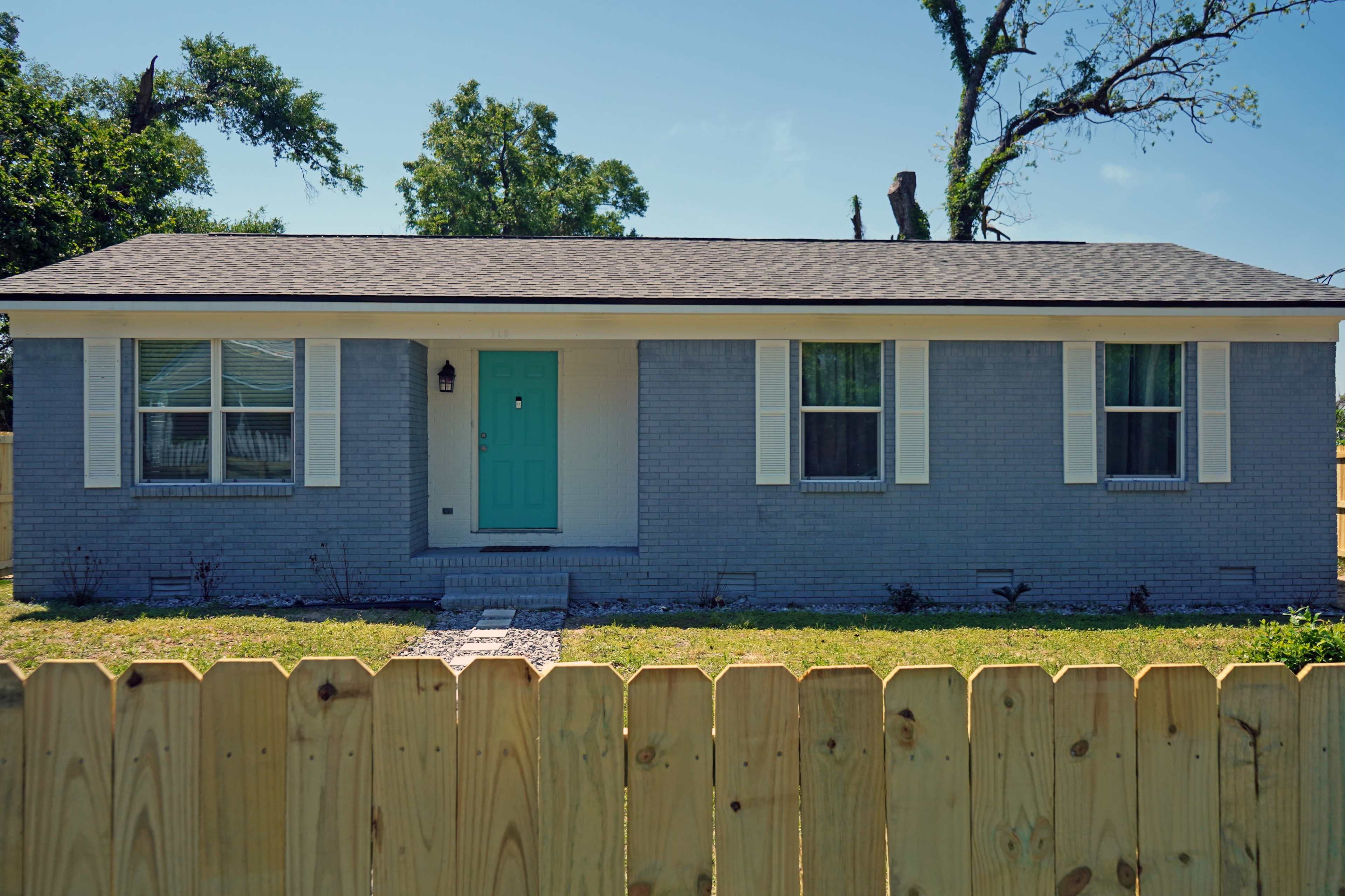 A single-story gray house with a turquoise front door and white shutters sits behind a wooden fence in a grassy yard.