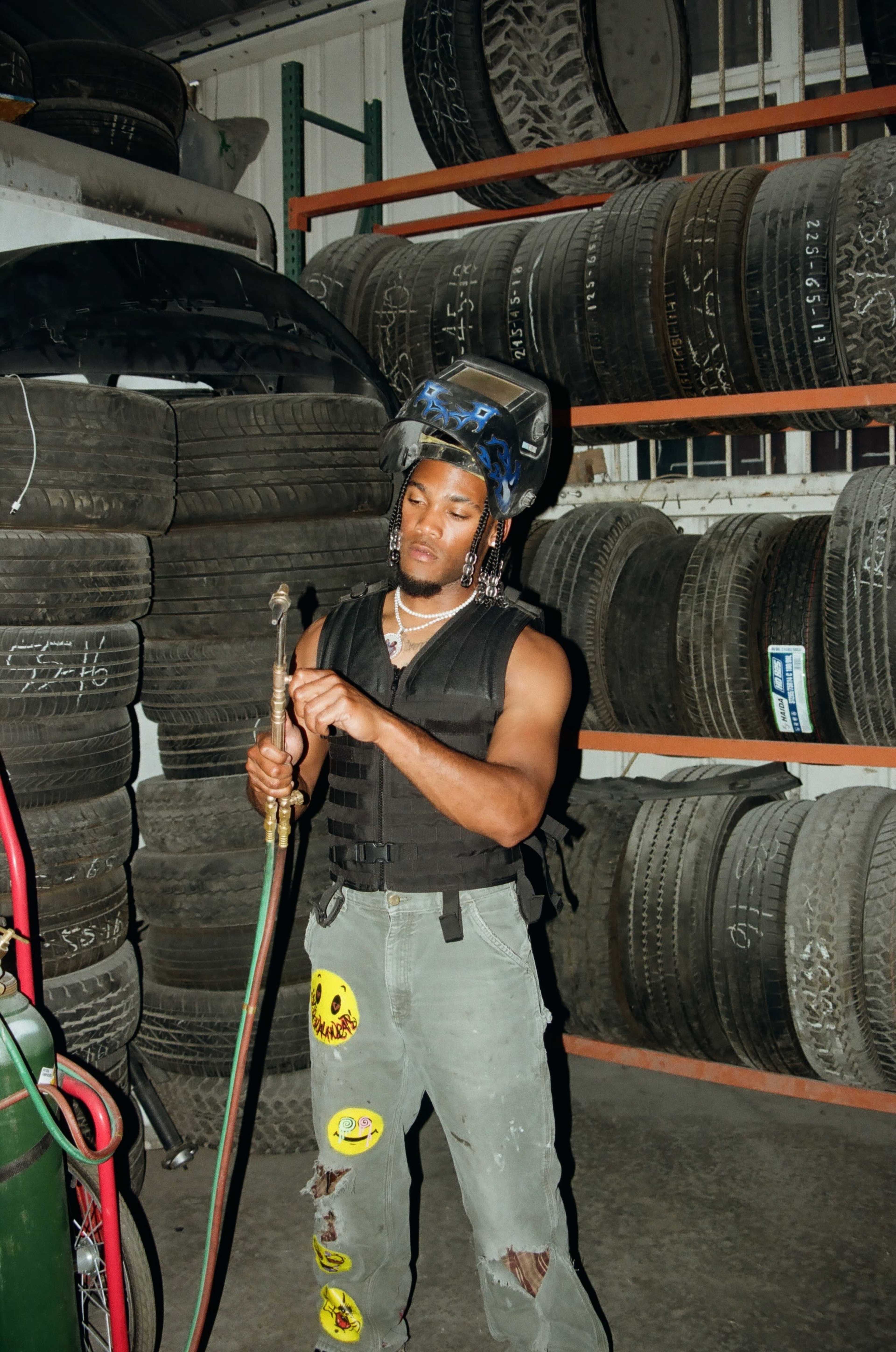 A person wears a protective helmet and holds a welding torch in a tire storage area filled with stacked tires.