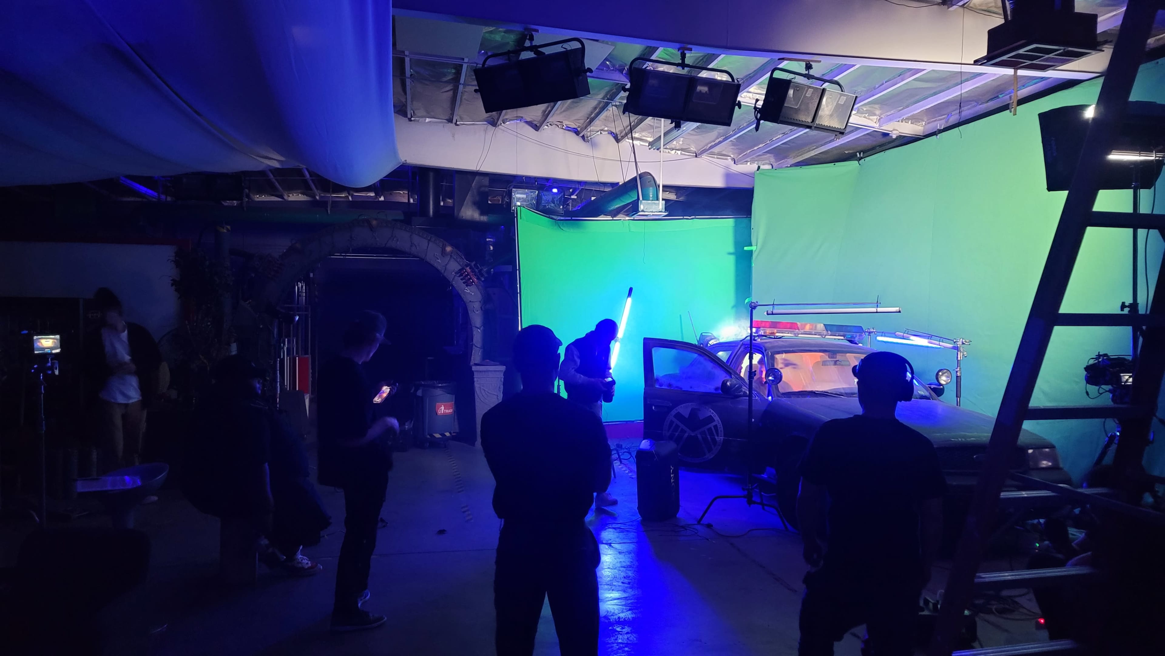 A film crew is working in a studio with a green screen, preparing equipment around a modified vehicle under artificial lighting.