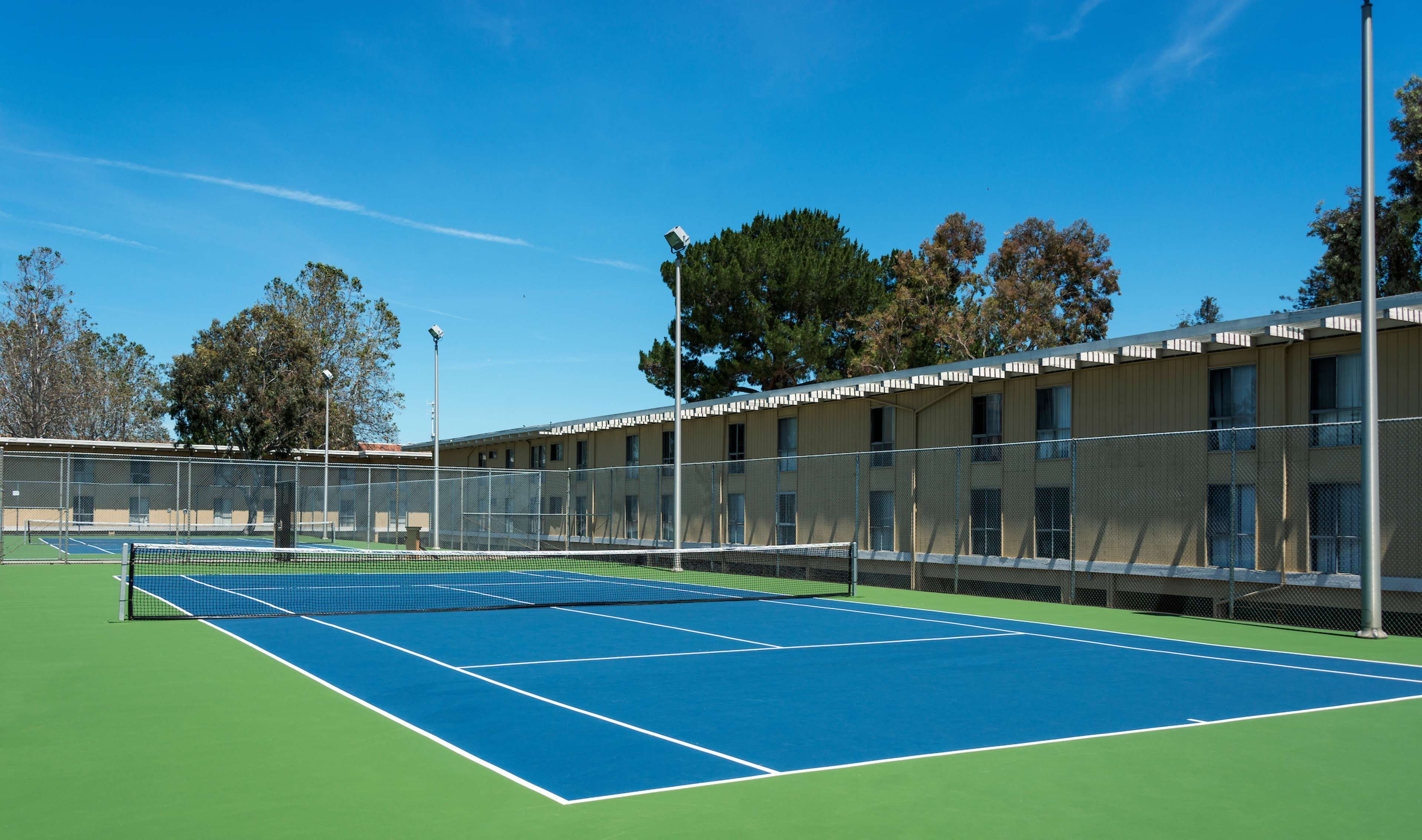 Several tennis courts with blue surfaces and a backdrop of a building and trees under a clear blue sky.