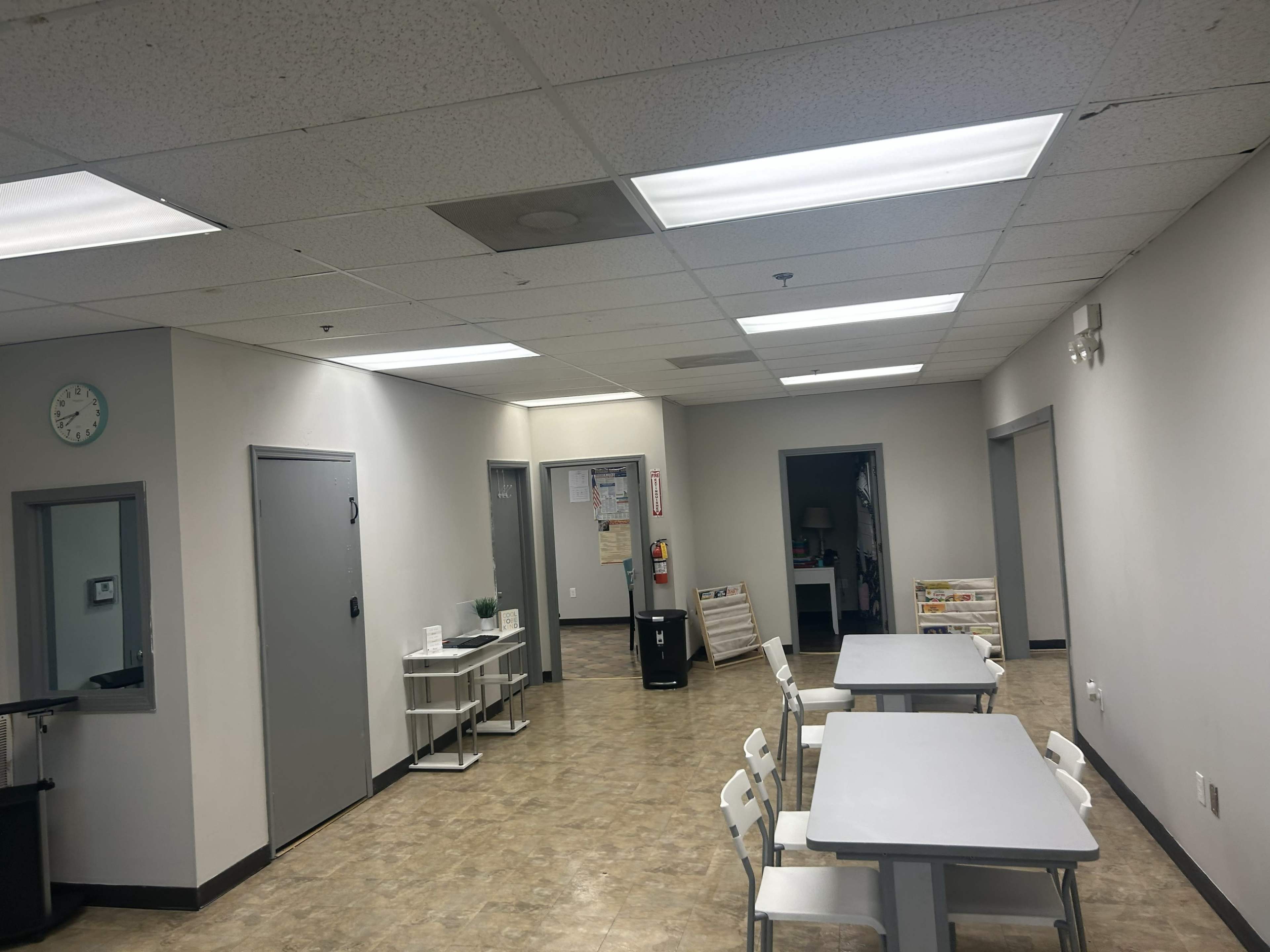 The image shows a spacious, brightly lit room with tables and chairs arranged in the center, flanked by doors leading to other areas.