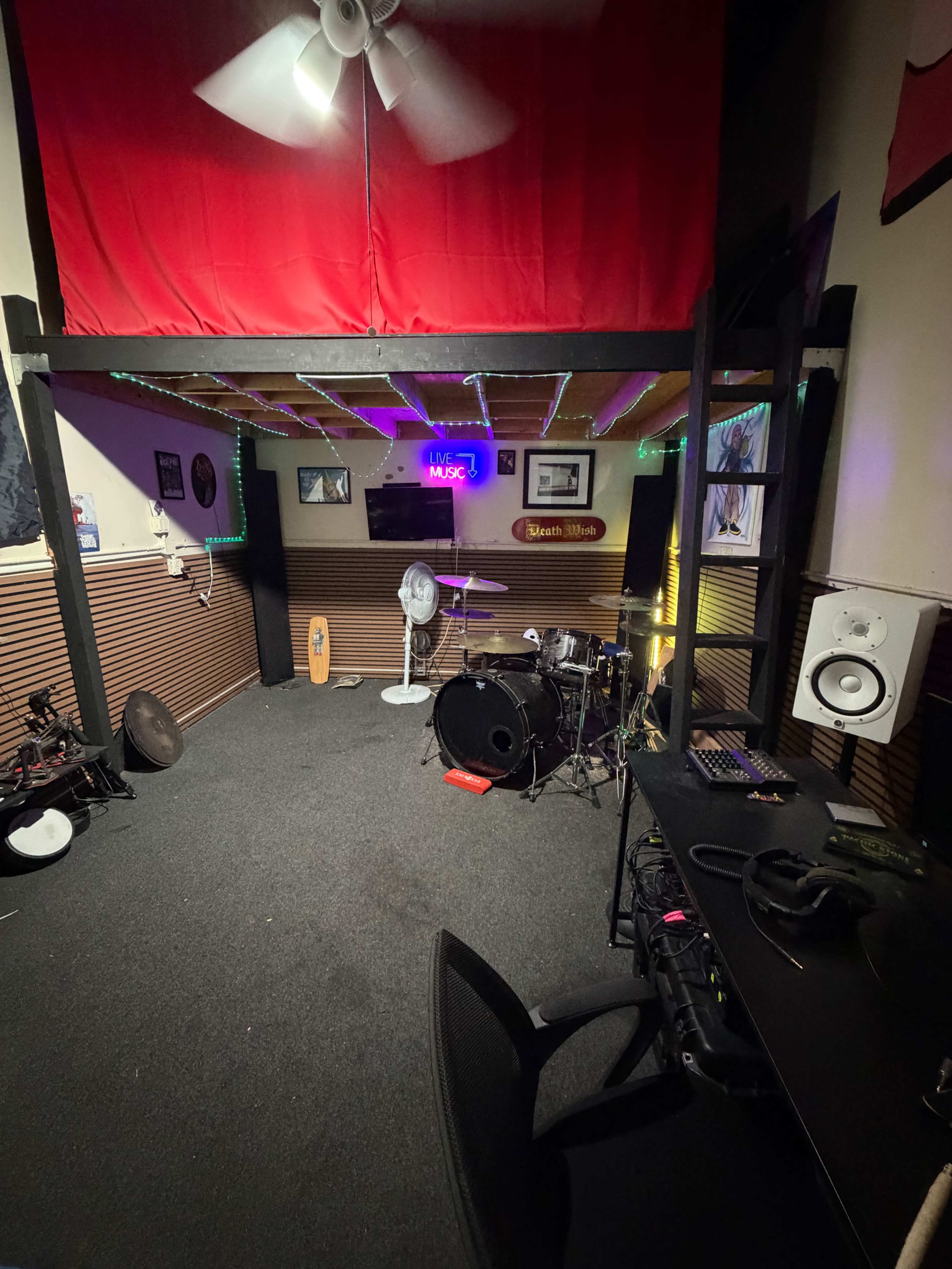 The image shows a music practice room featuring a drum set, a computer desk, and a large red curtain hanging above a lofted area.