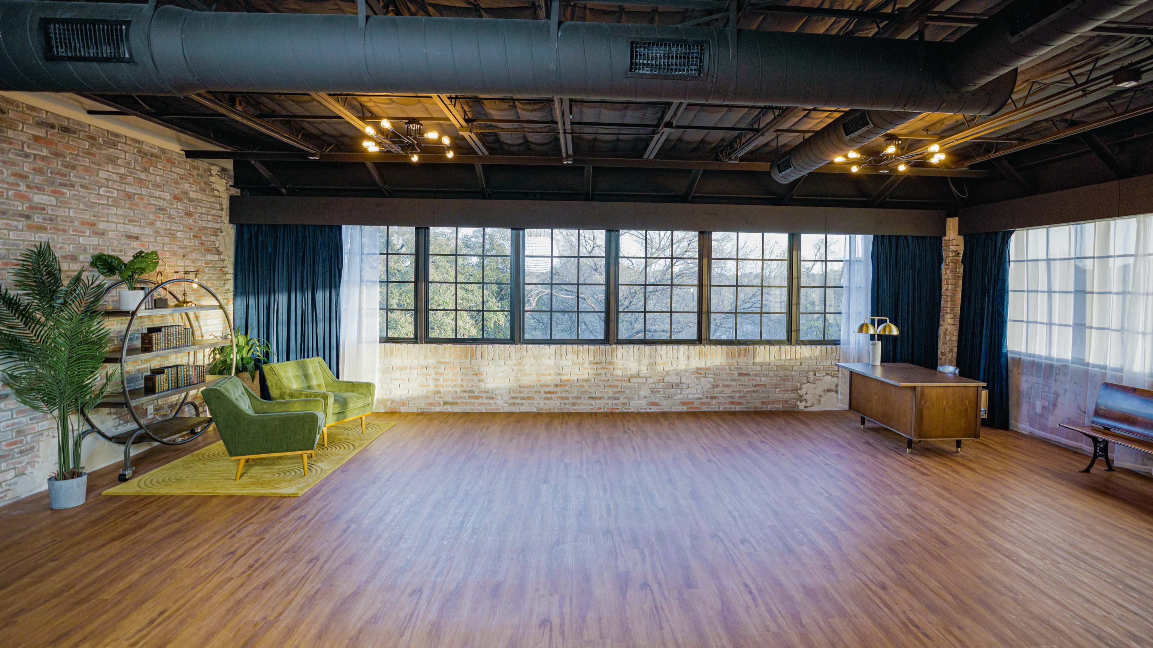 The image shows a spacious, modern room with large windows, wooden flooring, and minimal furniture, including two green chairs, a wooden desk, and a decorative plant.