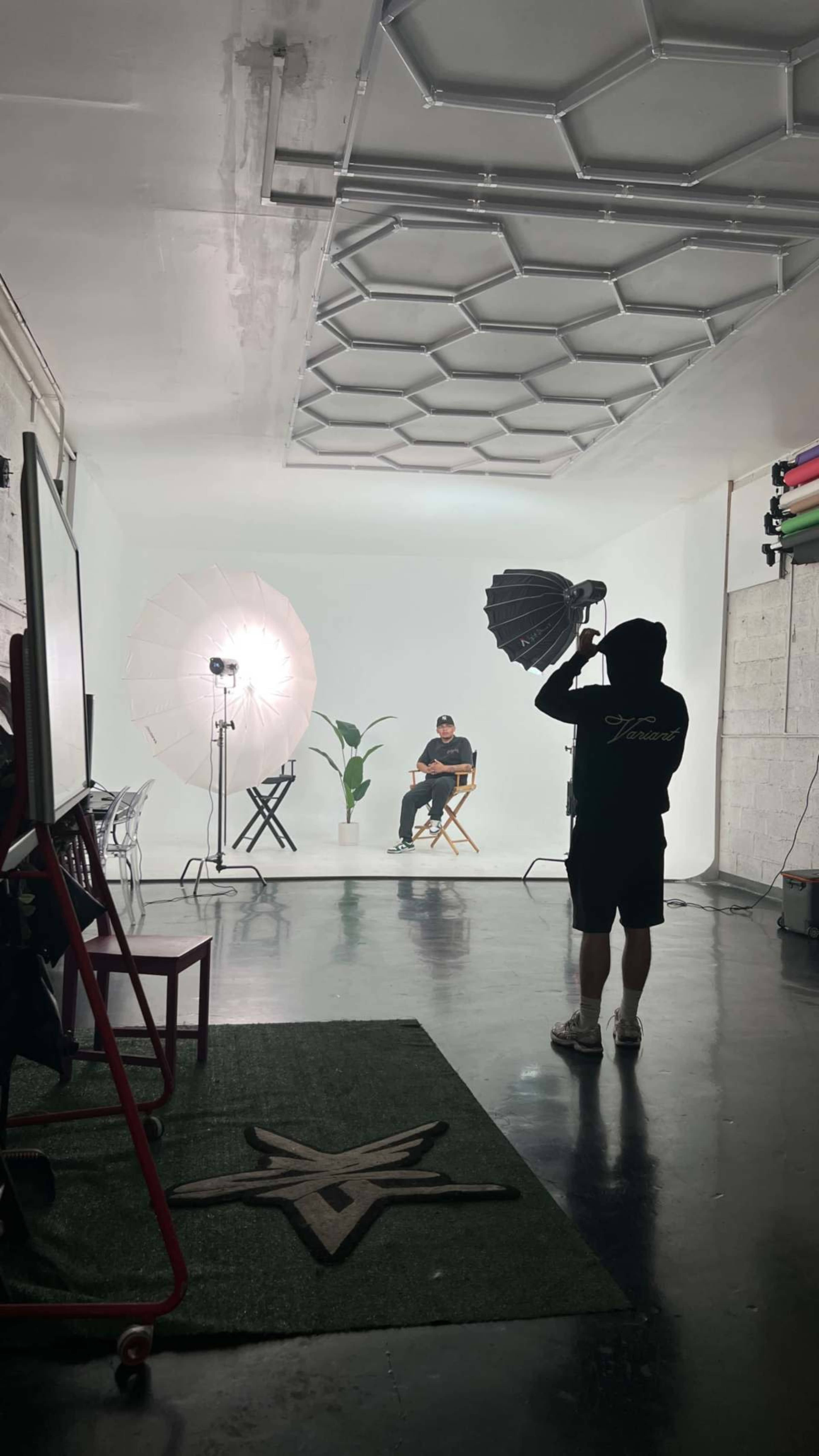 A photography studio with a person sitting on a chair surrounded by softbox lights and a plant, while another person sets up equipment in the foreground.