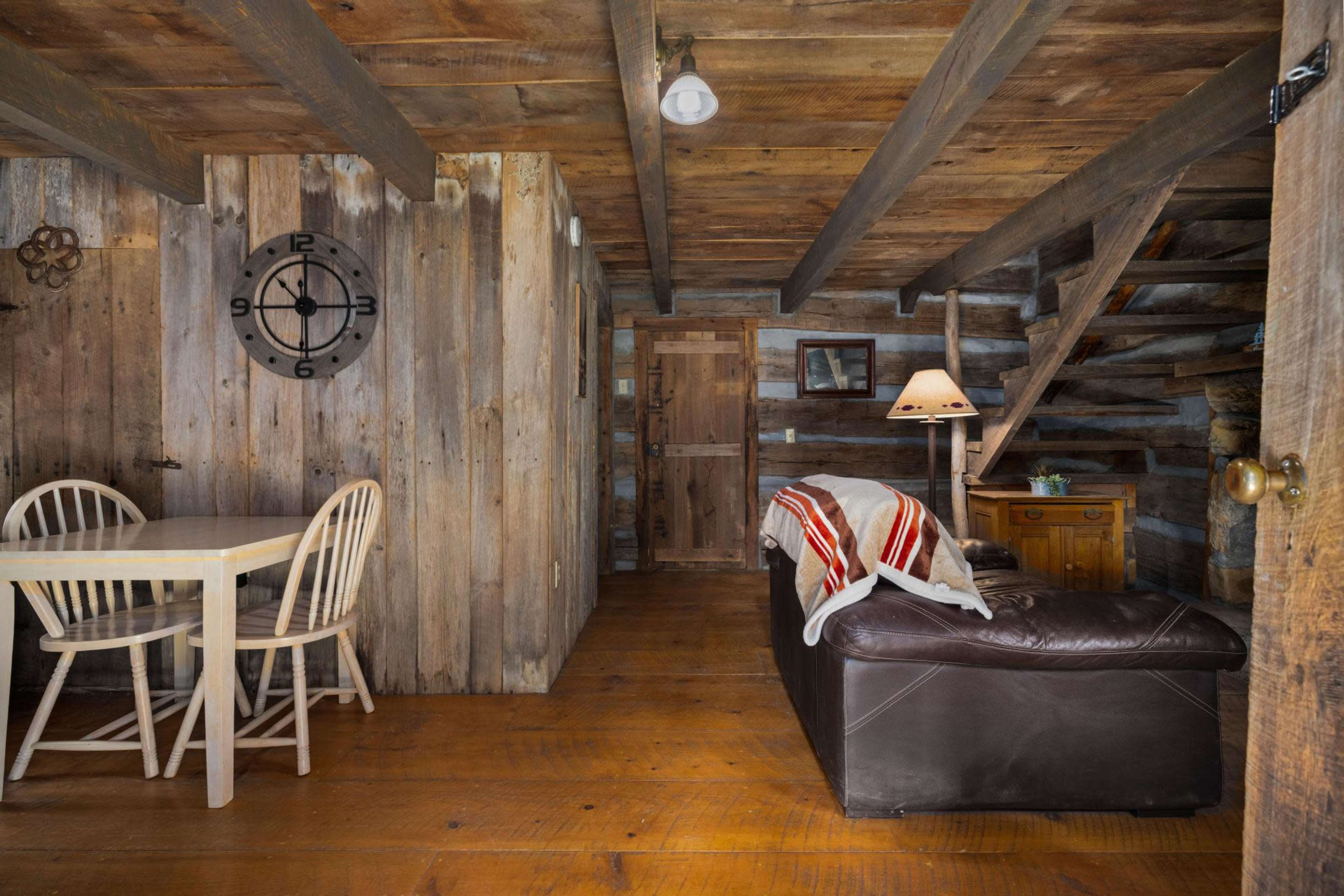 The image shows a rustic interior of a wooden cabin featuring a dining table with two chairs, a leather sofa, a floor lamp, and a staircase leading to an upper level.