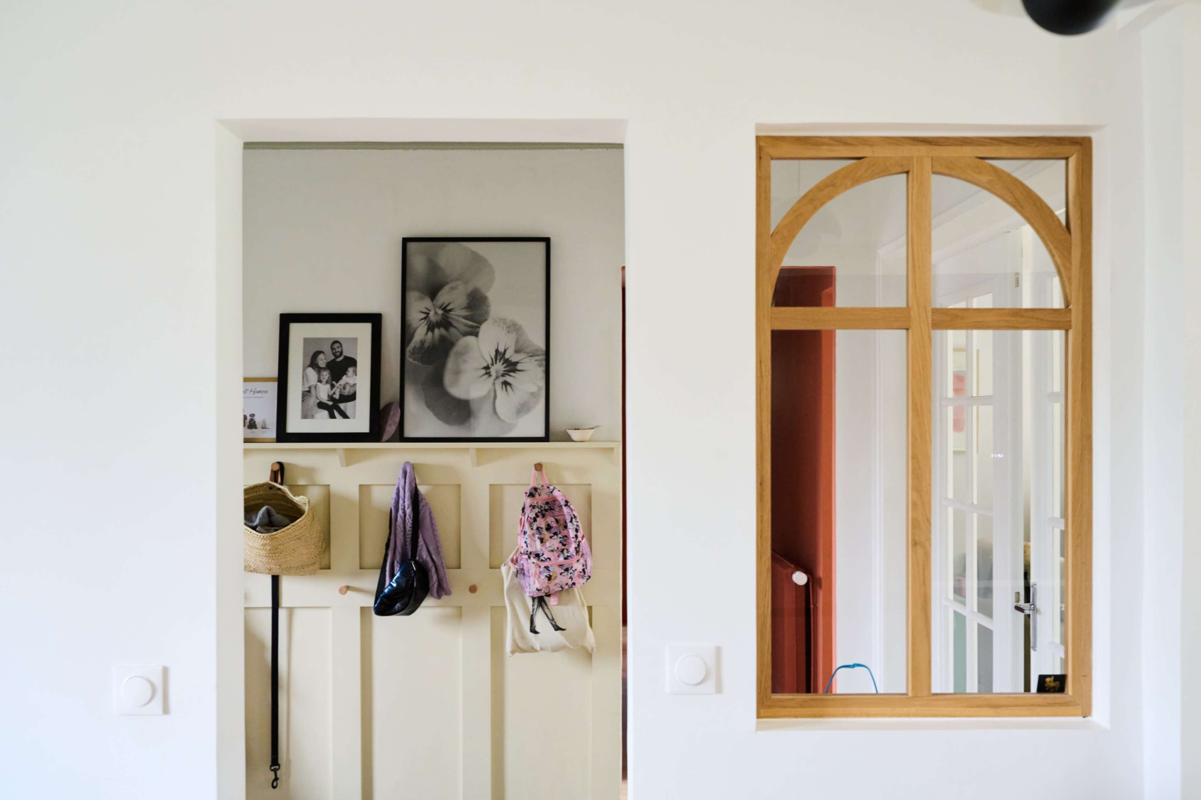 The image features a bright hallway with a coat rack on one side, framed photographs on the wall, and a large window-like opening with wooden trim on the opposite side.