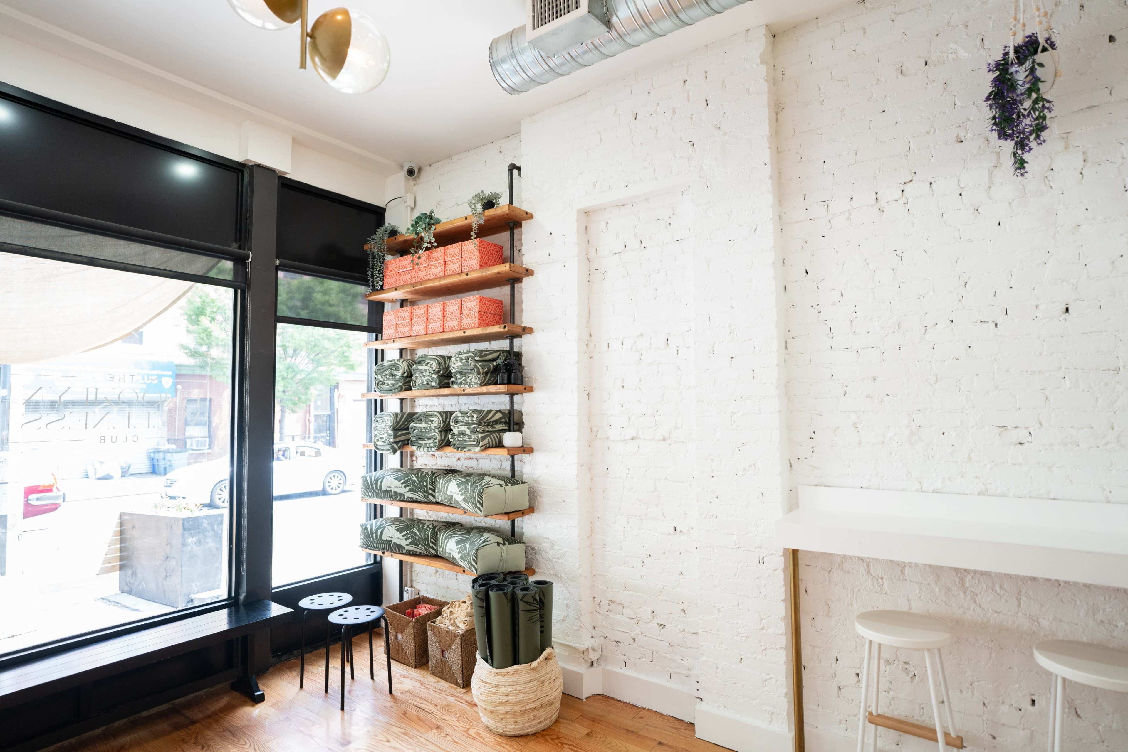 A minimalist interior featuring a white brick wall with wooden shelves displaying folded textiles and potted plants, alongside a seating area by a large window.