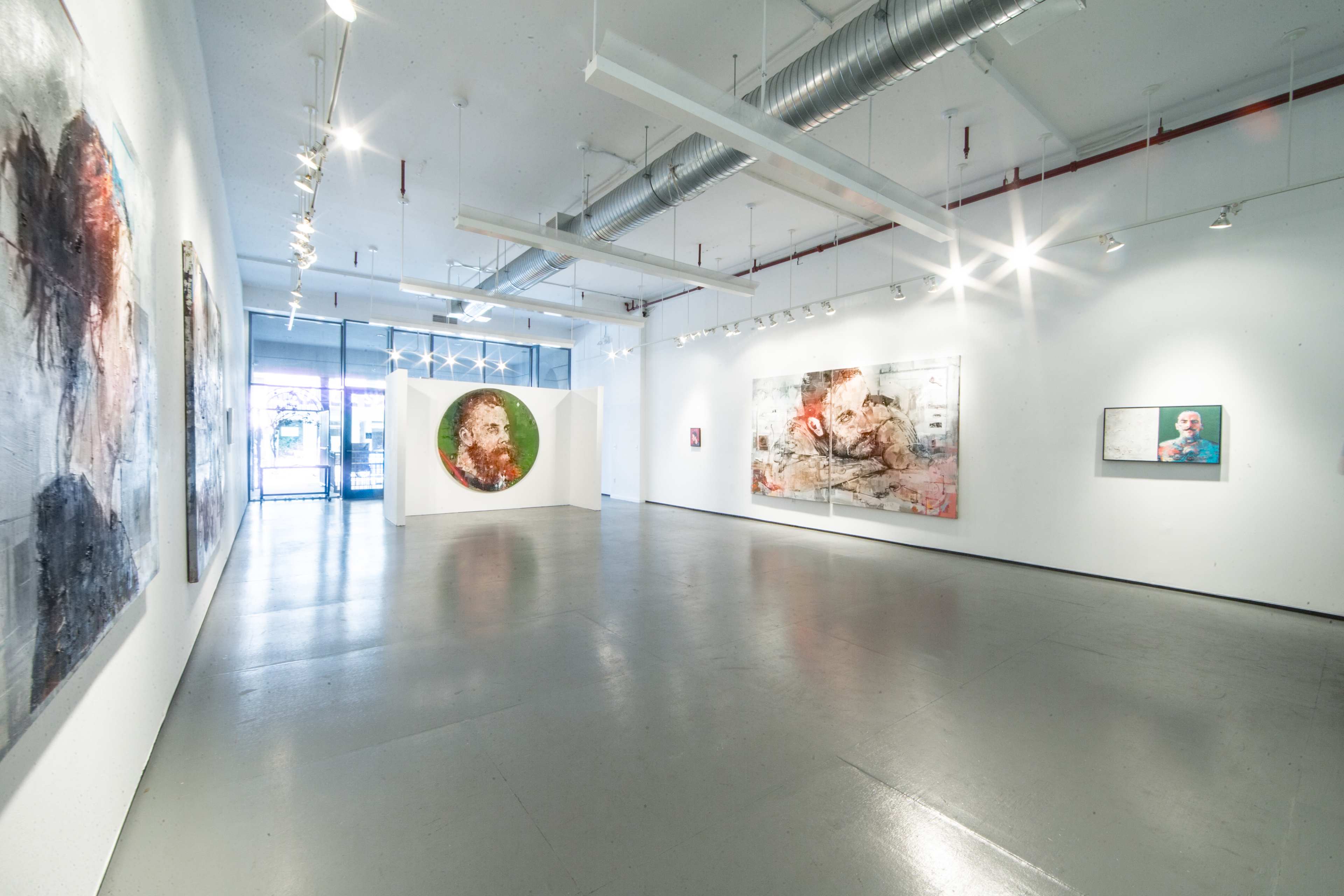 An art gallery features several large abstract paintings on white walls and a polished concrete floor, with natural light coming in from large windows.