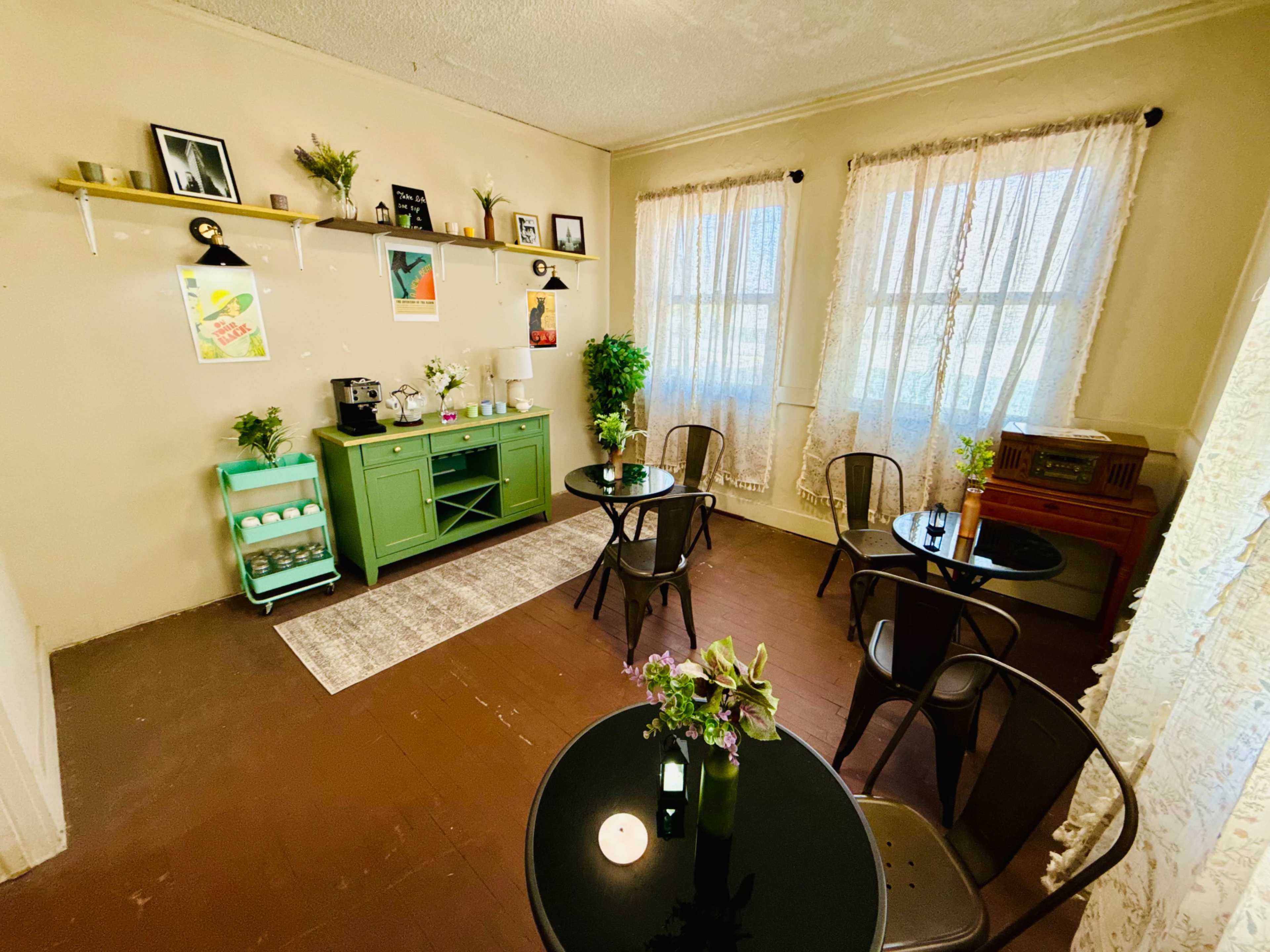 The image shows a cozy room with two small round tables, green furniture, and large windows dressed with light curtains, adorned with various decorative items and plants.