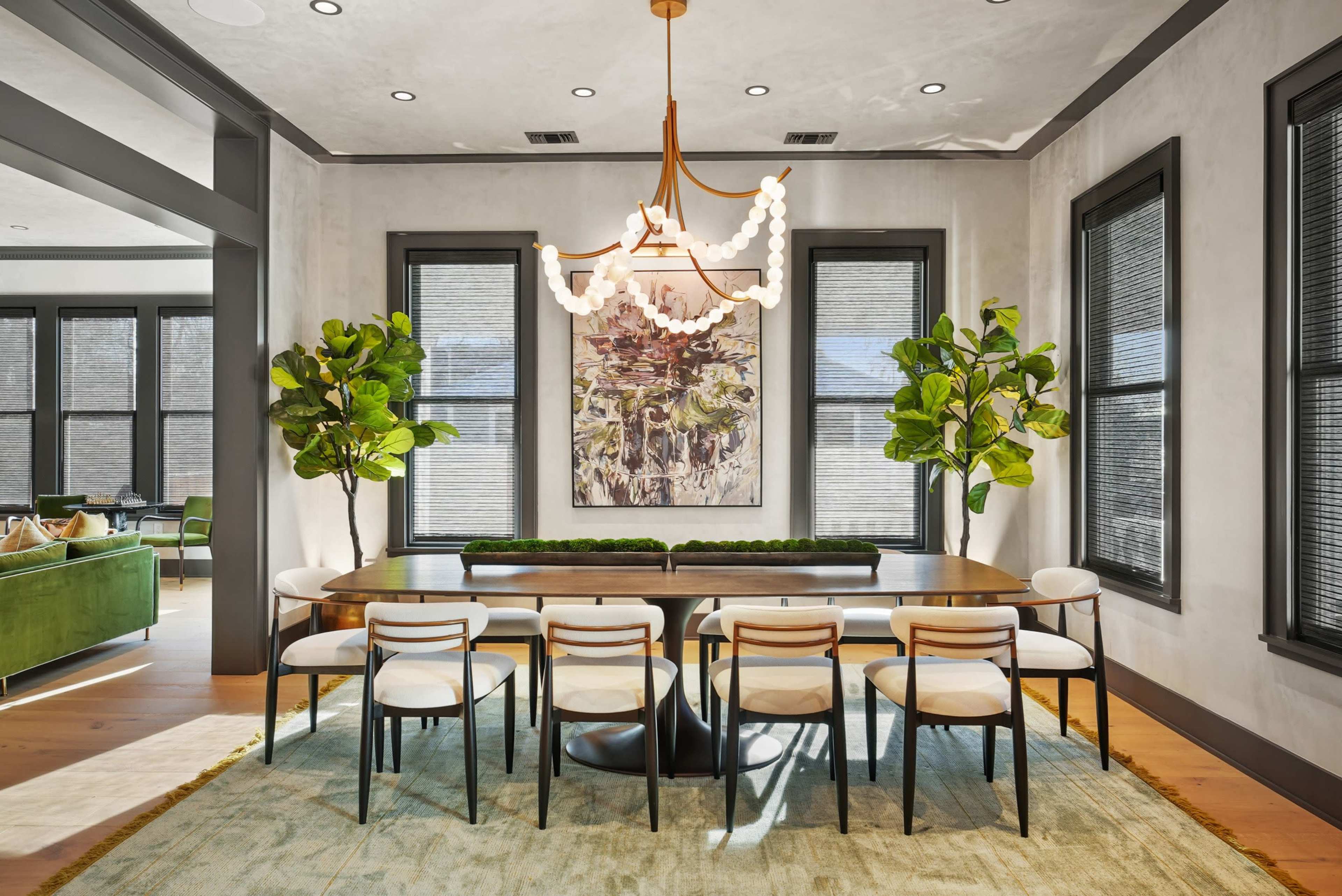 A modern dining room features a long wooden table surrounded by eight chairs, with a chandelier and potted plants enhancing the decor.