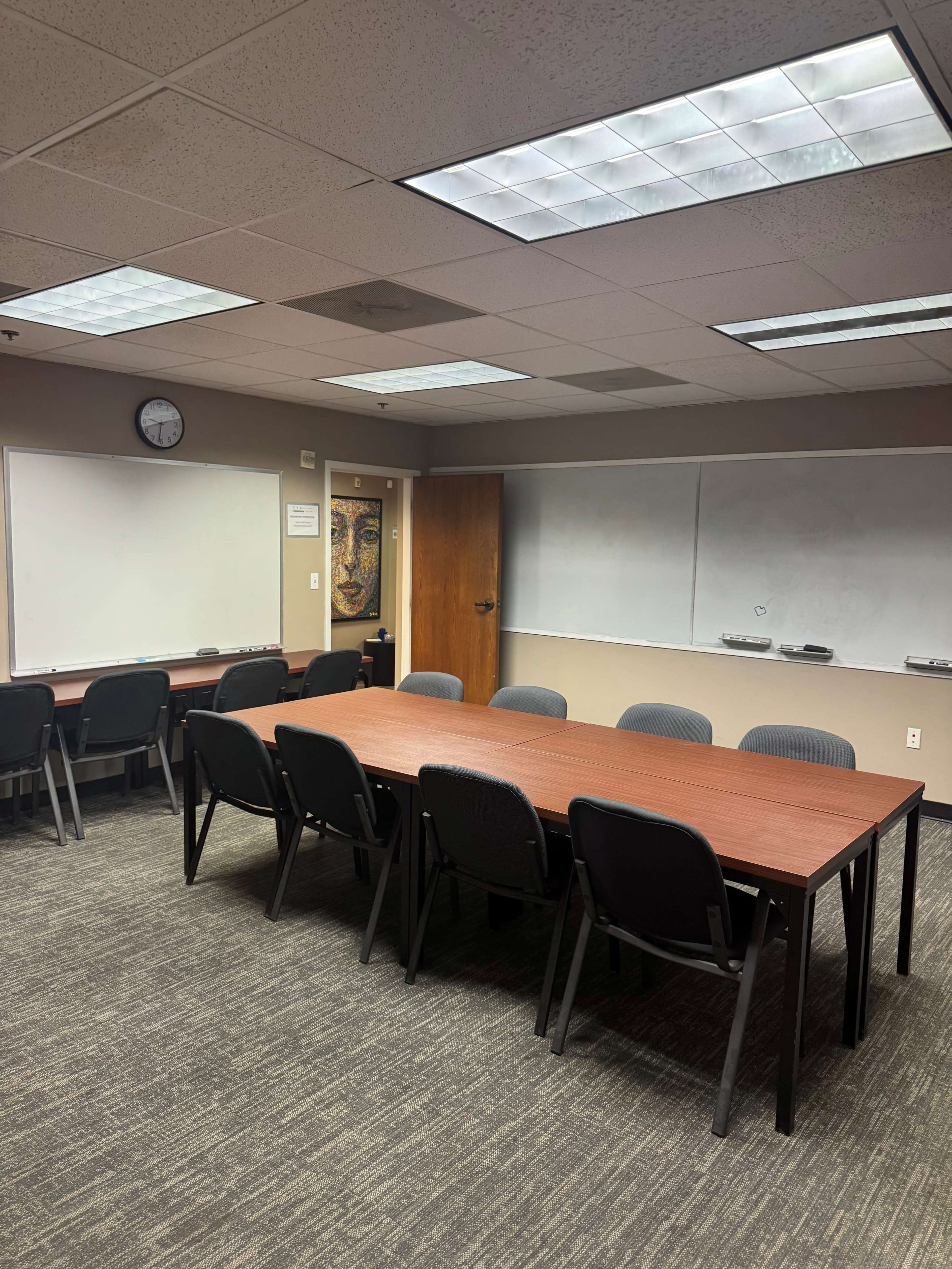 The image shows a conference room with a long wooden table surrounded by black chairs, whiteboards on two walls, and a clock on the back wall.