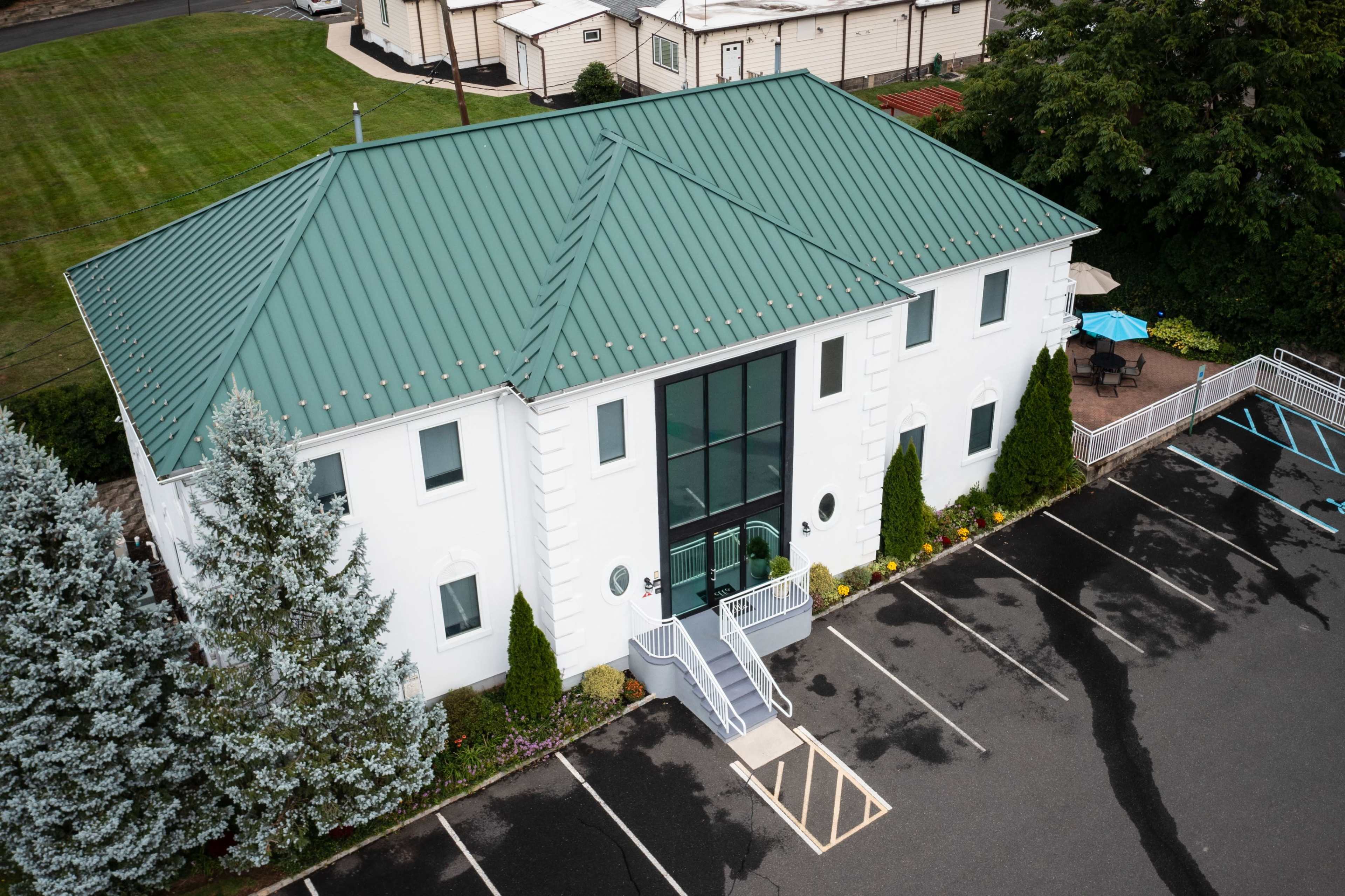A large white building with a green metal roof features a central glass entrance and is surrounded by landscaped grass and parking spaces.