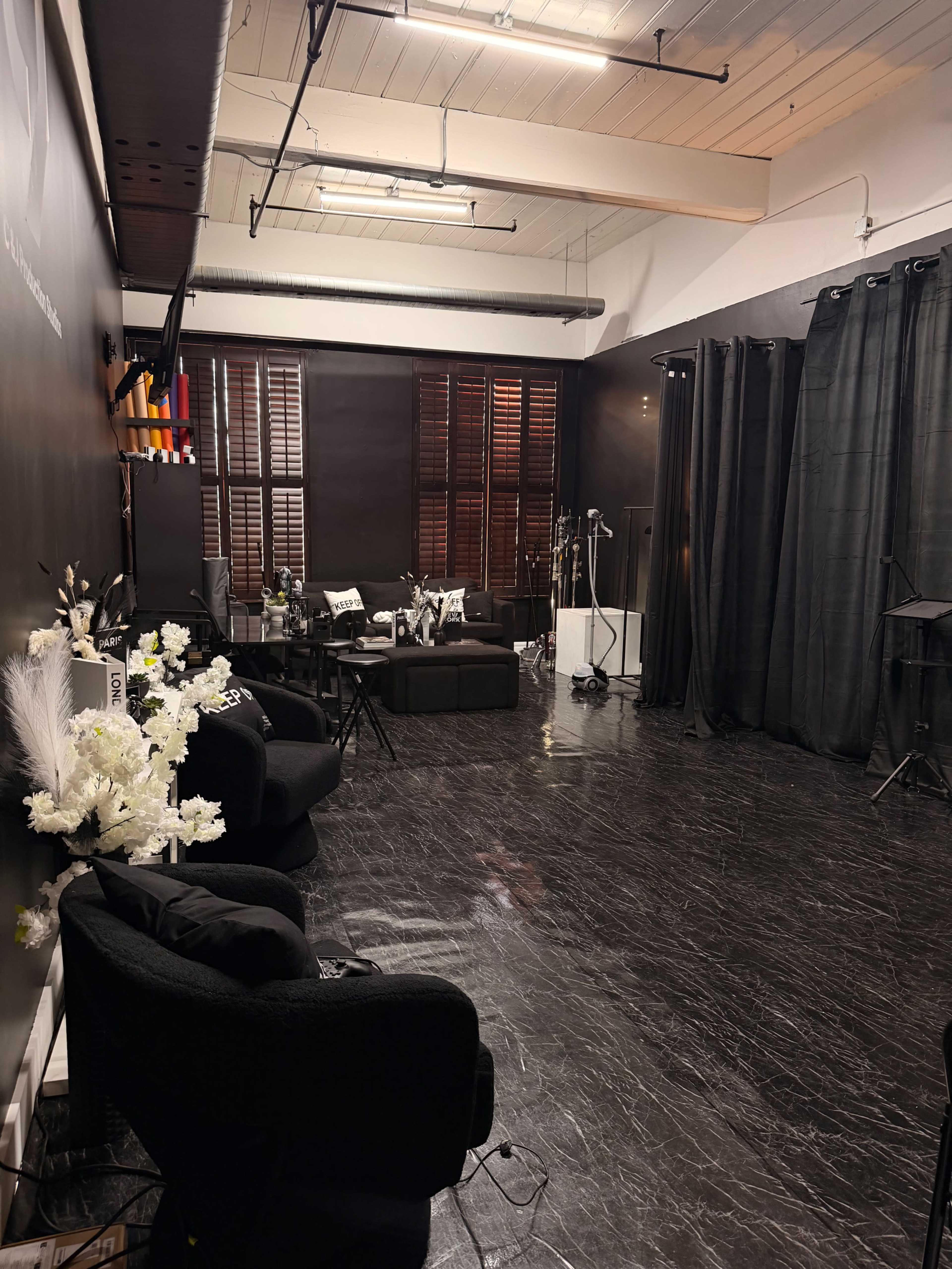 The image shows a dimly lit studio space with black walls, sleek flooring, and various pieces of furniture including sofas and chairs, accompanied by decorative elements like plants and photography equipment.