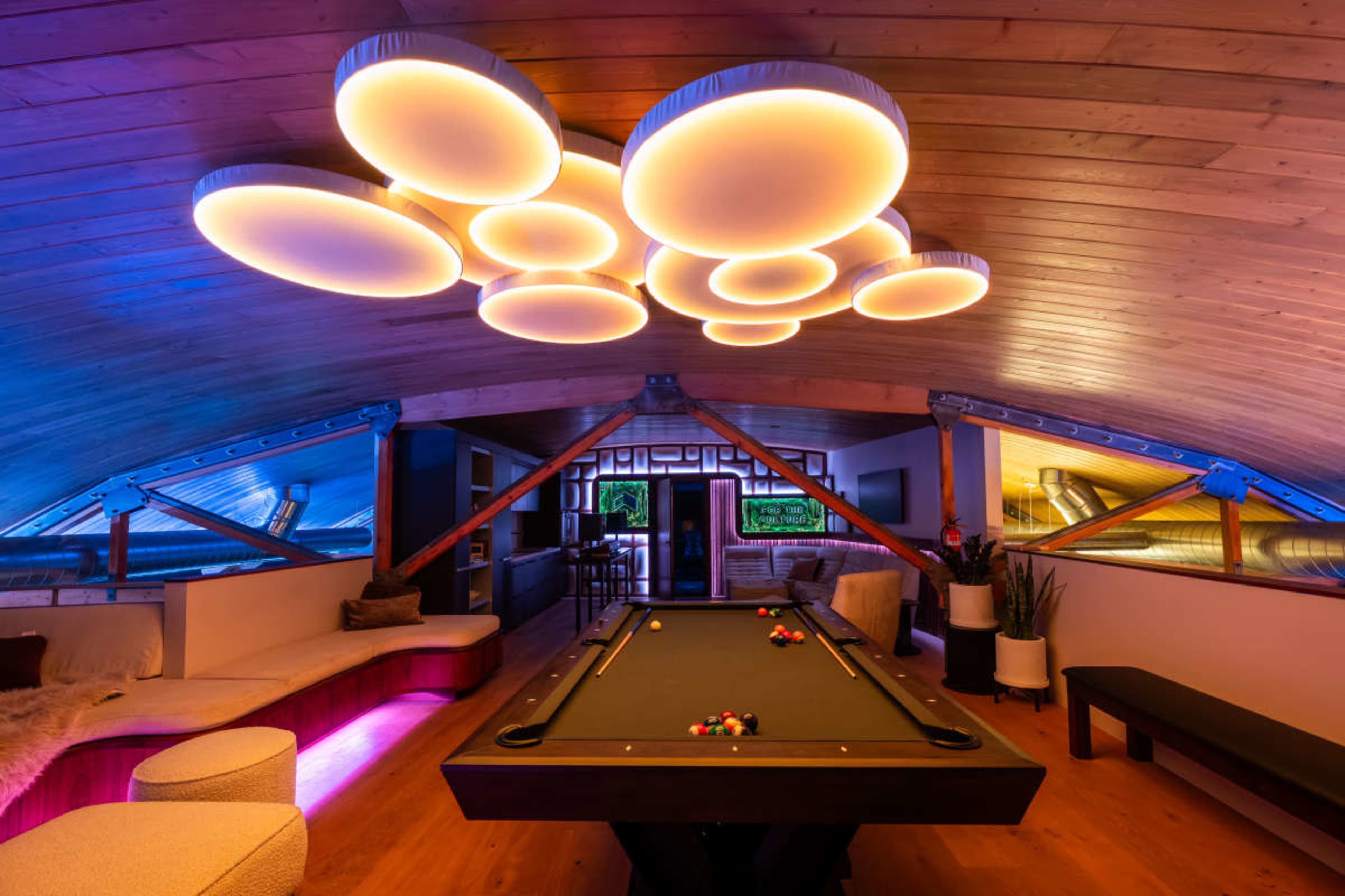 The image shows a modern game room with a stylish pool table, illuminated by unique circular ceiling lights, and featuring a cozy seating area.