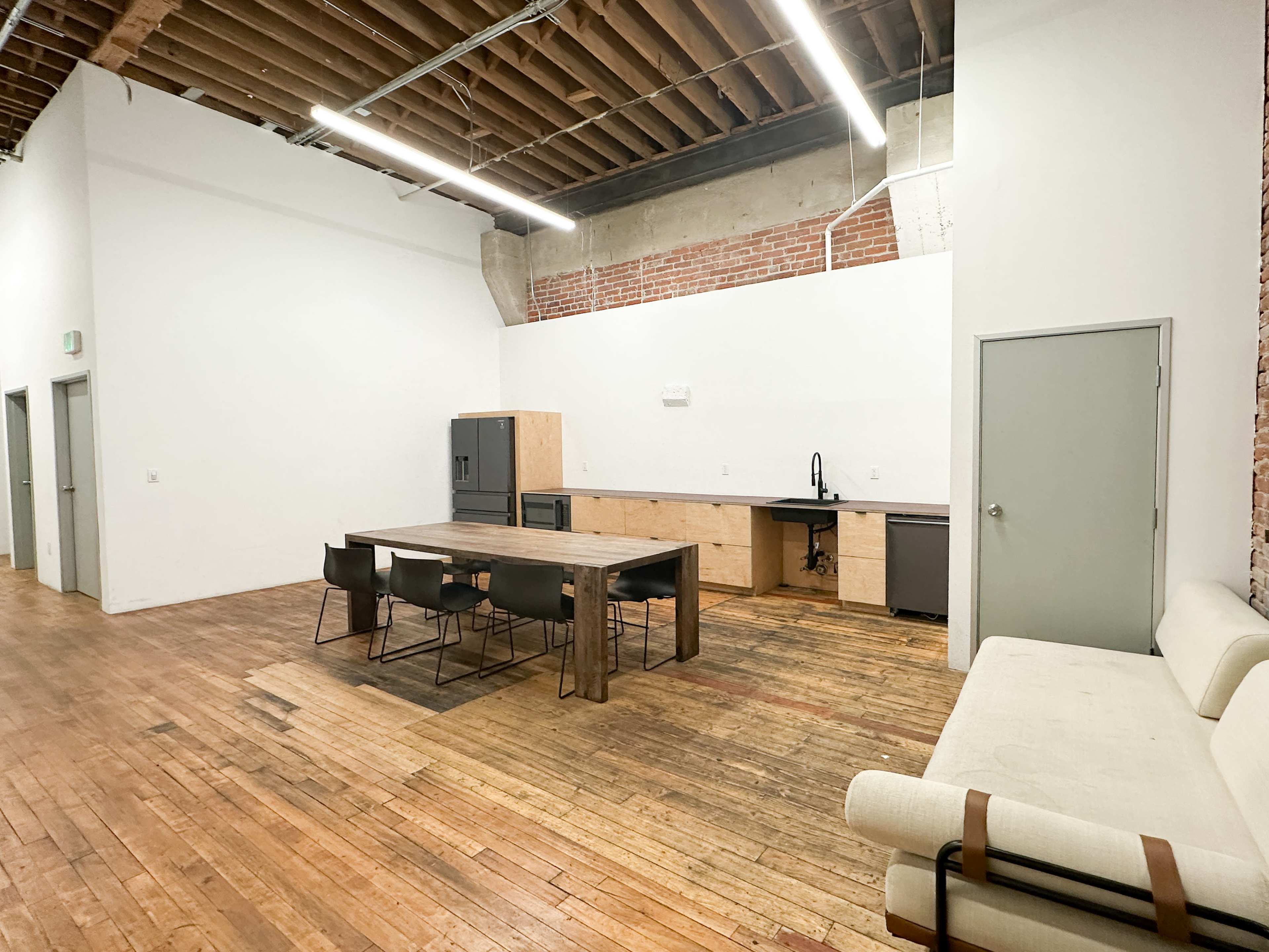 The image shows a spacious, minimalistic kitchen and dining area with wooden flooring, a long table with chairs, a refrigerator, and modern appliances against white and brick walls.