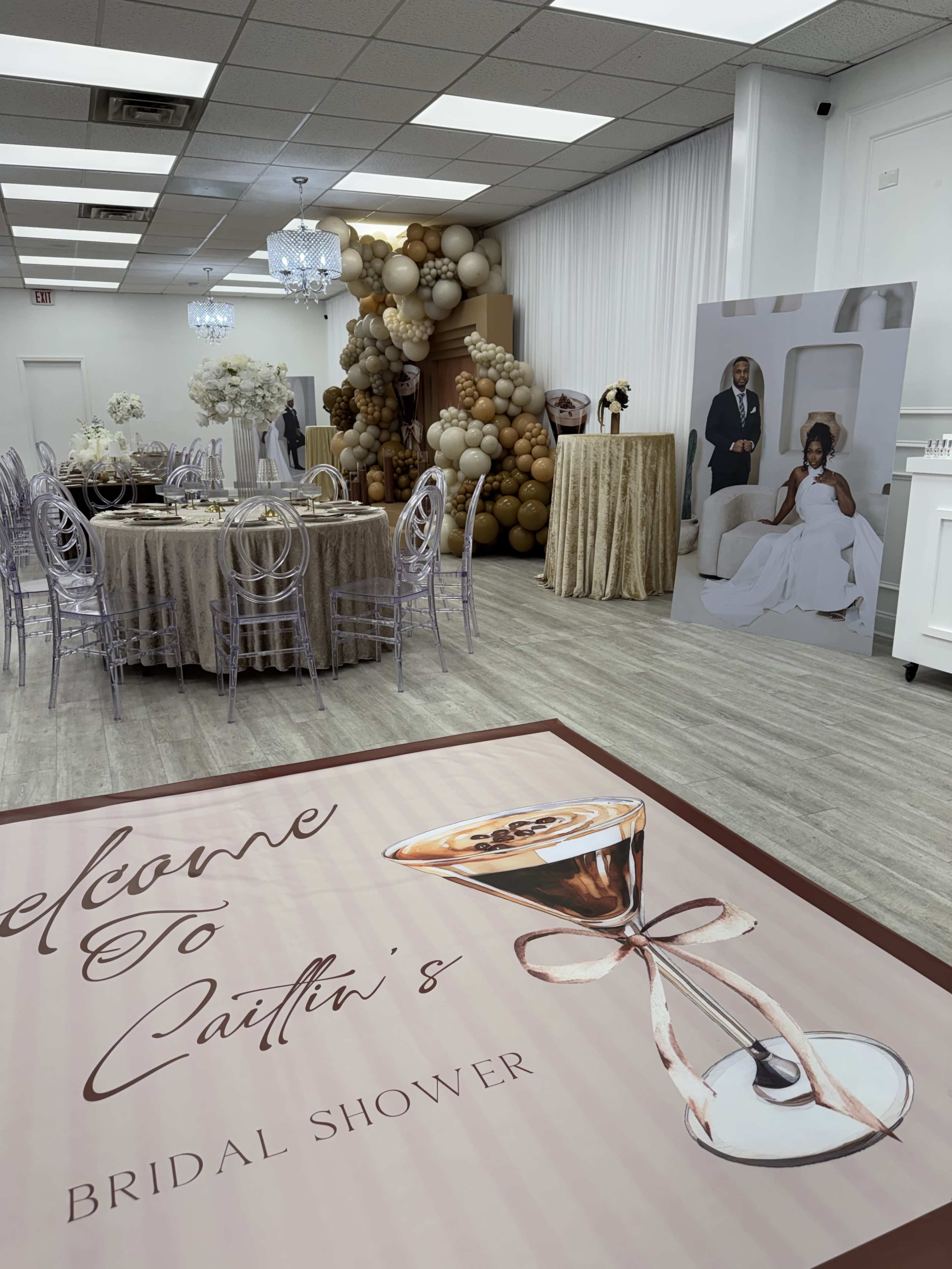 The image shows a decorated event space set up for a bridal shower, featuring a table with clear chairs, a balloon backdrop, and a welcome sign on the floor.