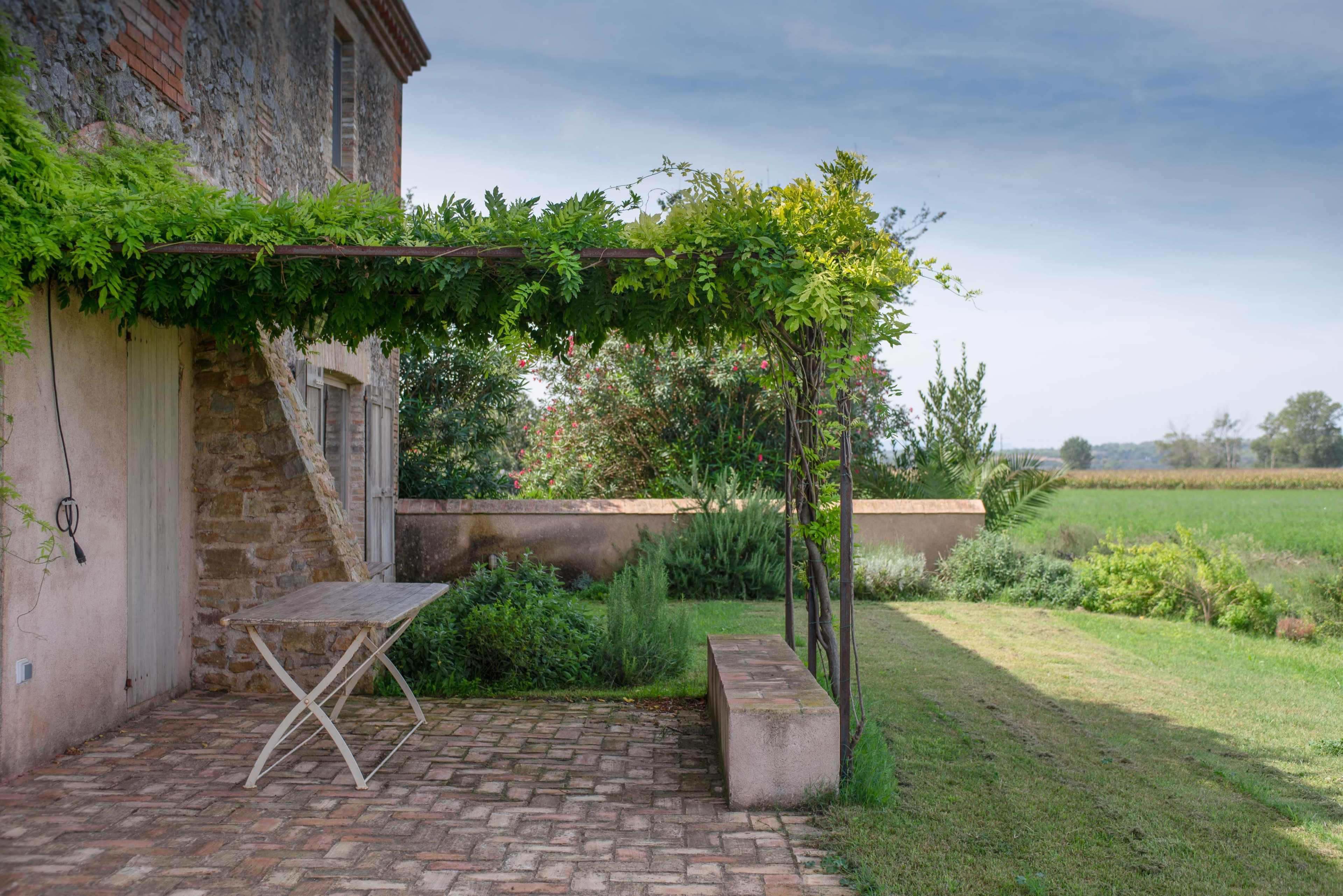 A stone farmhouse features a covered patio with a wooden table and a grassy area overlooking a field.
