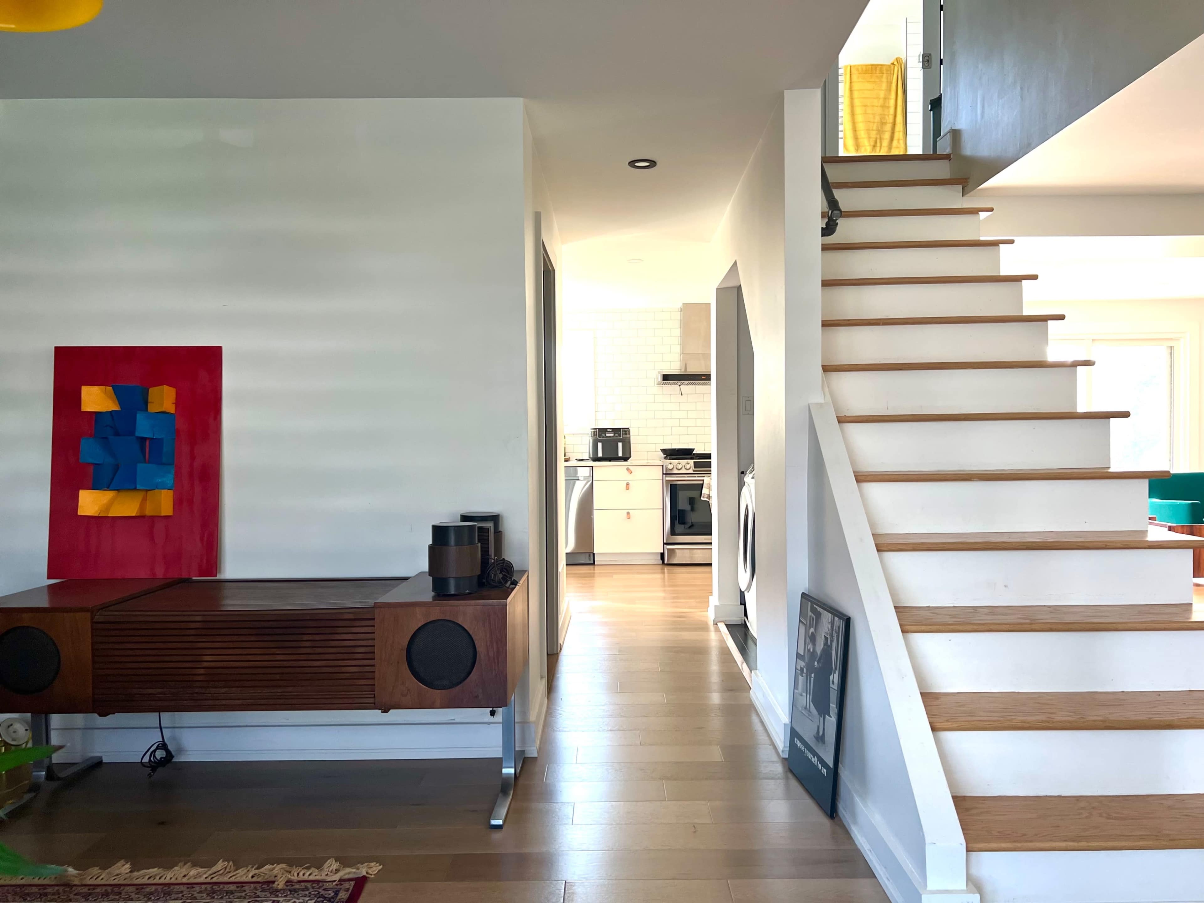 A modern interior with a staircase on the right, leading to an upper level, and a glimpse of a kitchen area in the background, with colorful artwork on the wall.