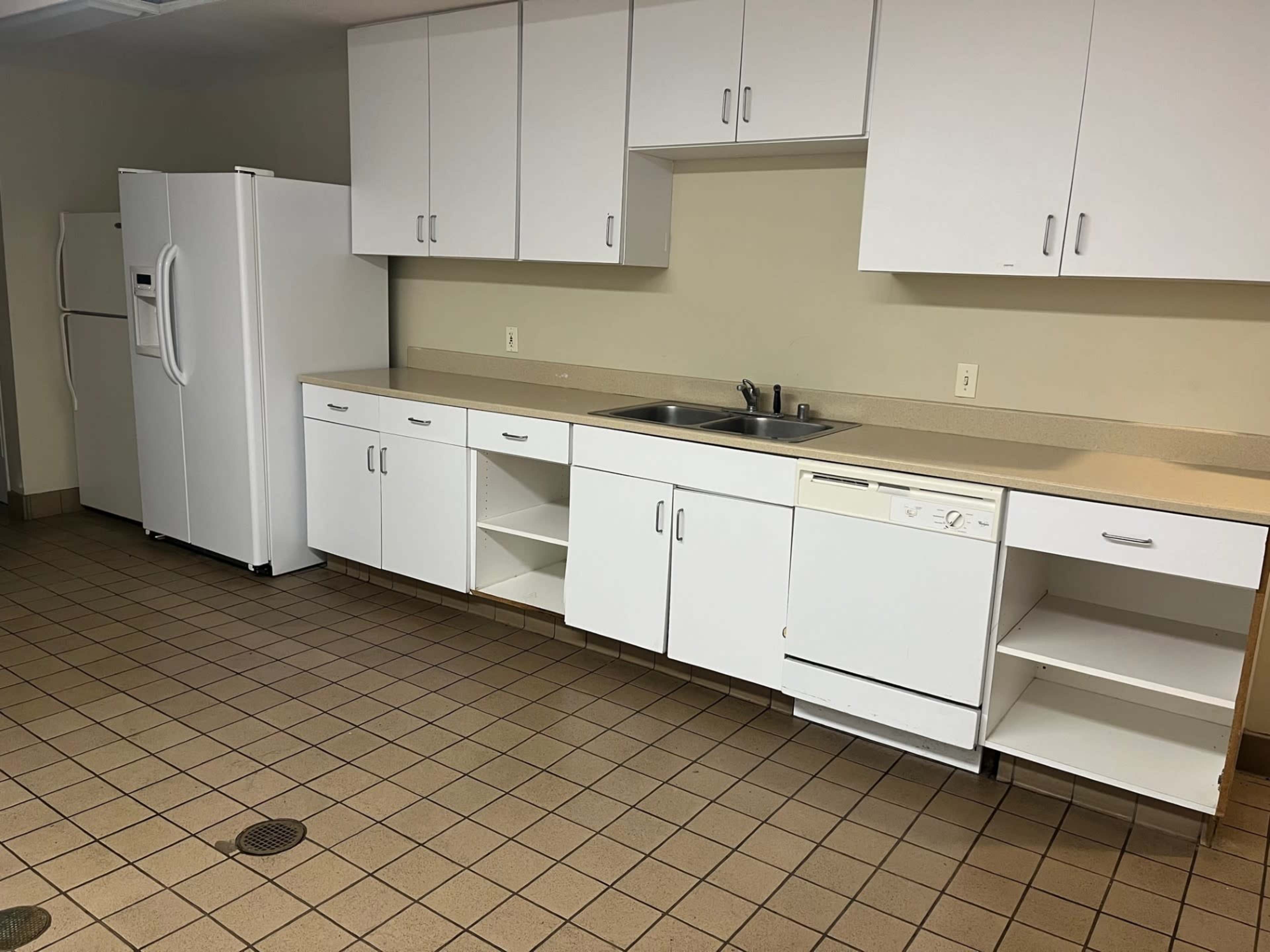 The image shows a kitchen interior with white cabinets, a double sink, a dishwasher, and a large refrigerator against a tiled floor.