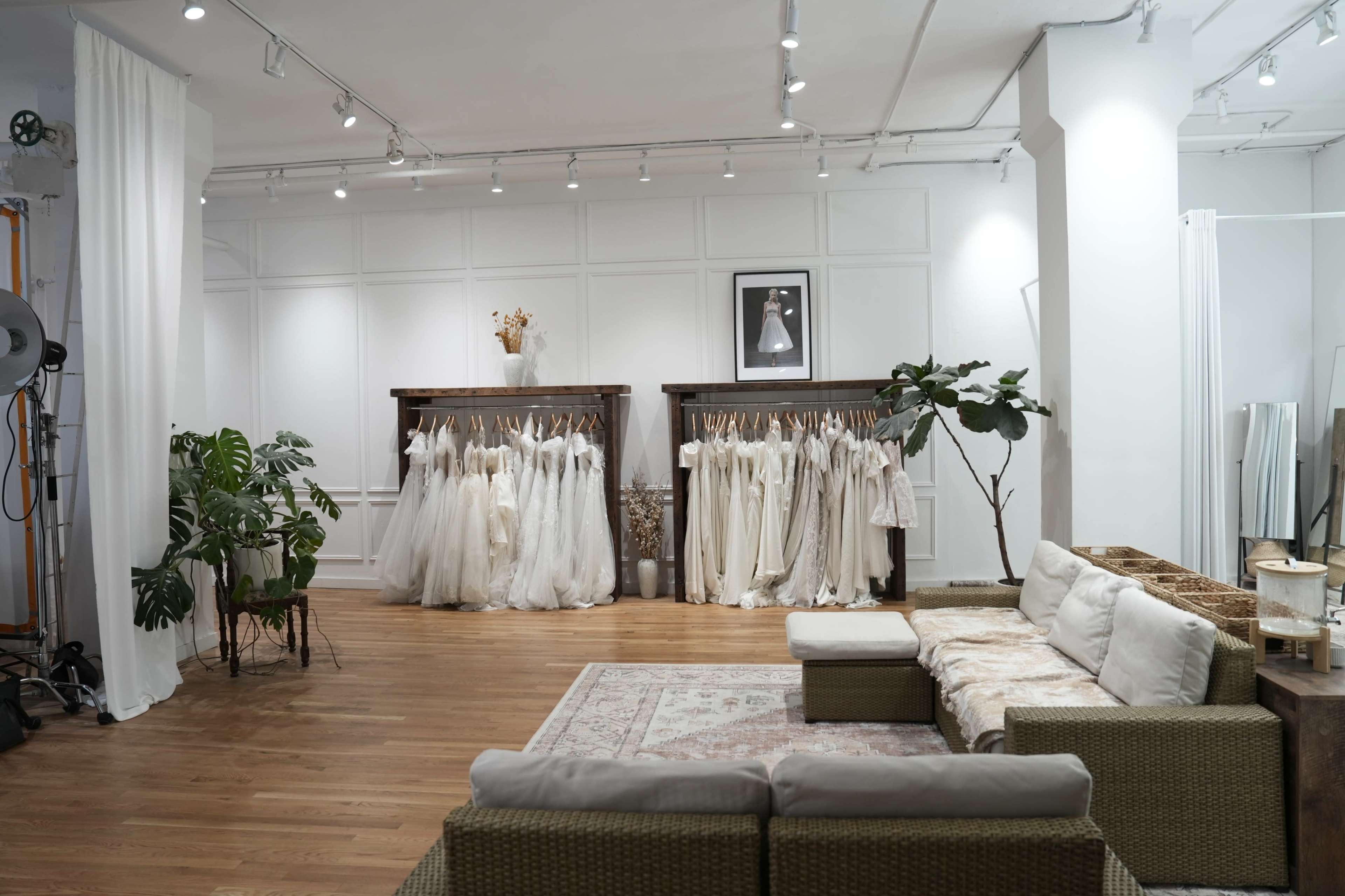 A spacious bridal boutique features racks of wedding dresses, a comfortable seating area, and plants, all set against white walls and hardwood flooring.