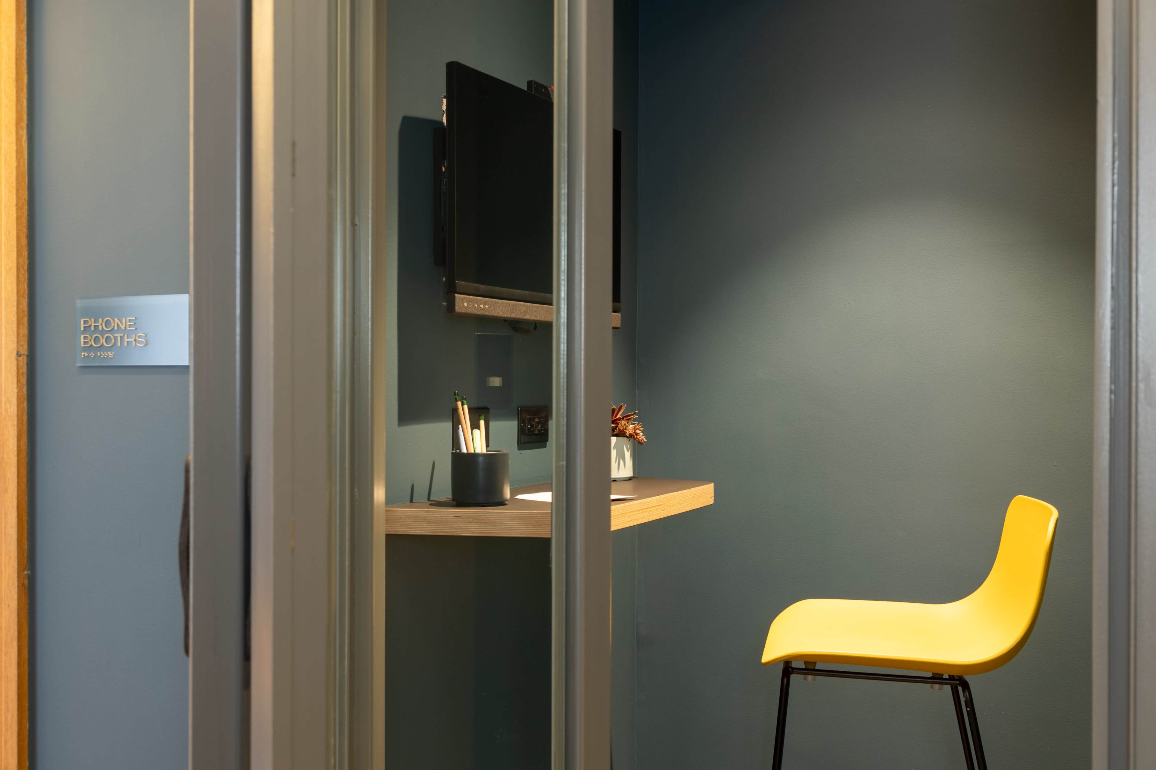 The image shows a small phone booth featuring a yellow chair, a wall-mounted television, and a wooden shelf with stationery against a dark blue wall.