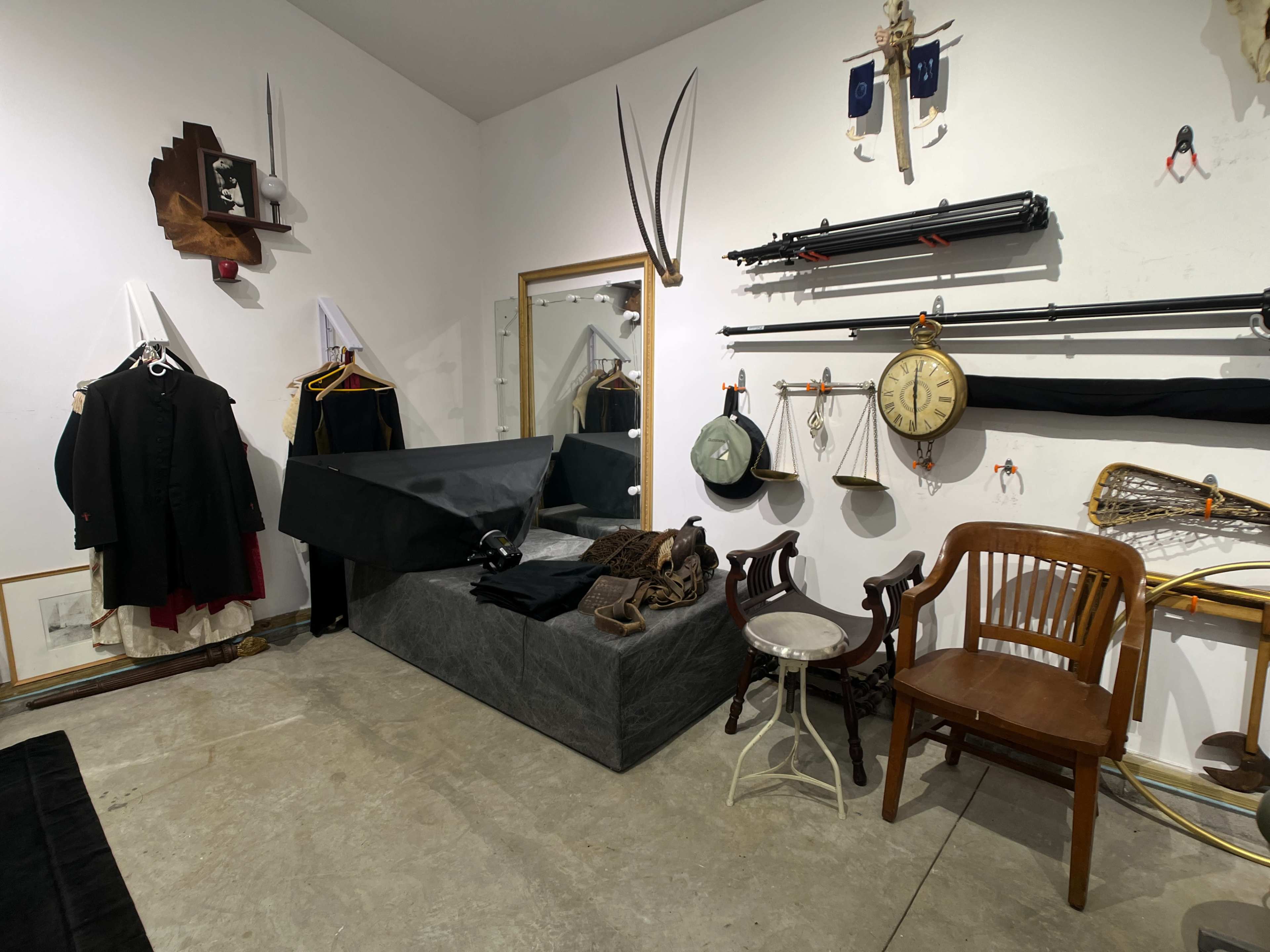 A room with various items, including clothing on hangers, vintage tools, a clock, and a wooden chair arranged around a central black-covered object.