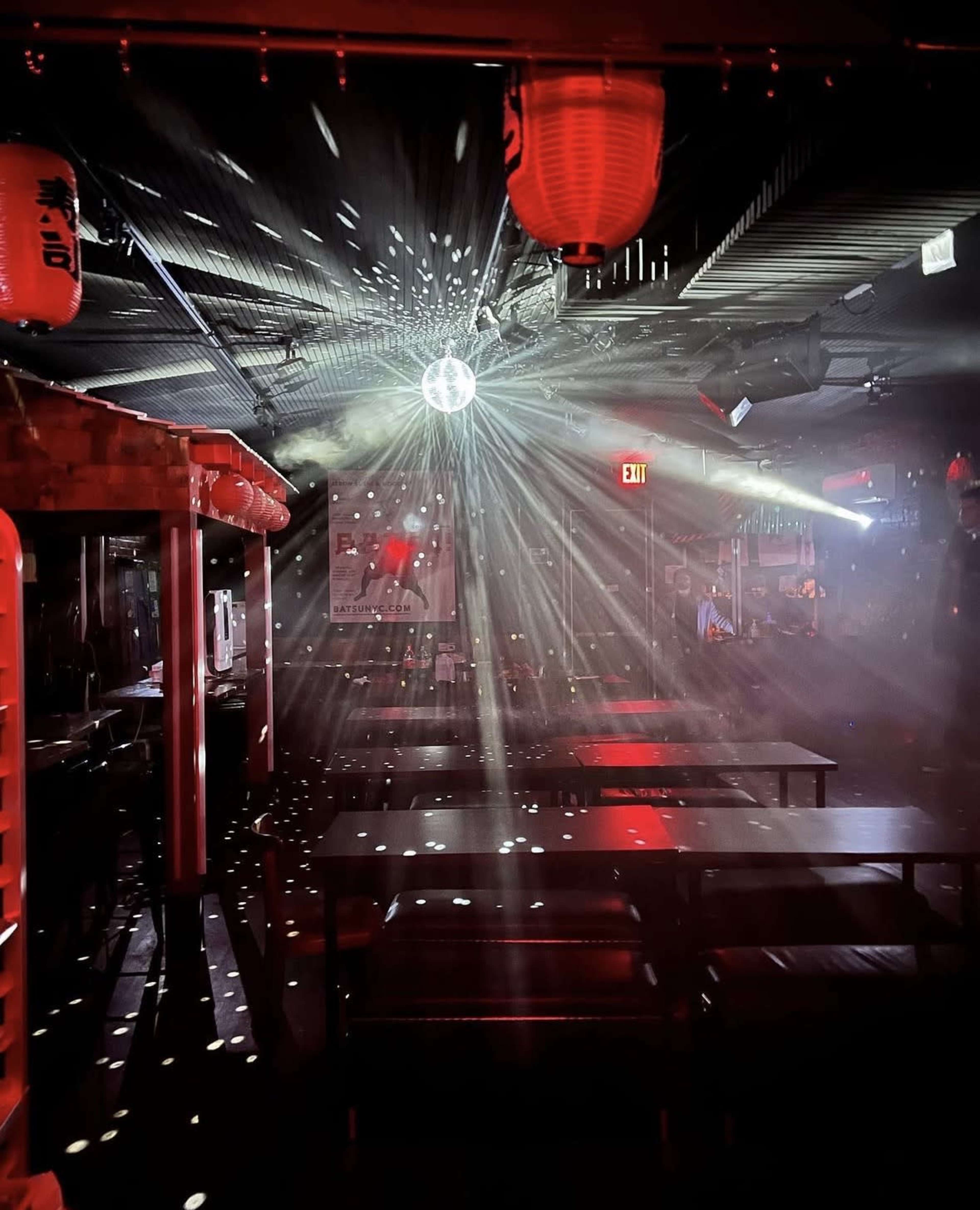 The interior of a dimly lit restaurant or bar features a disco ball reflecting light beams, with red lanterns hanging from the ceiling and rows of tables arranged beneath.