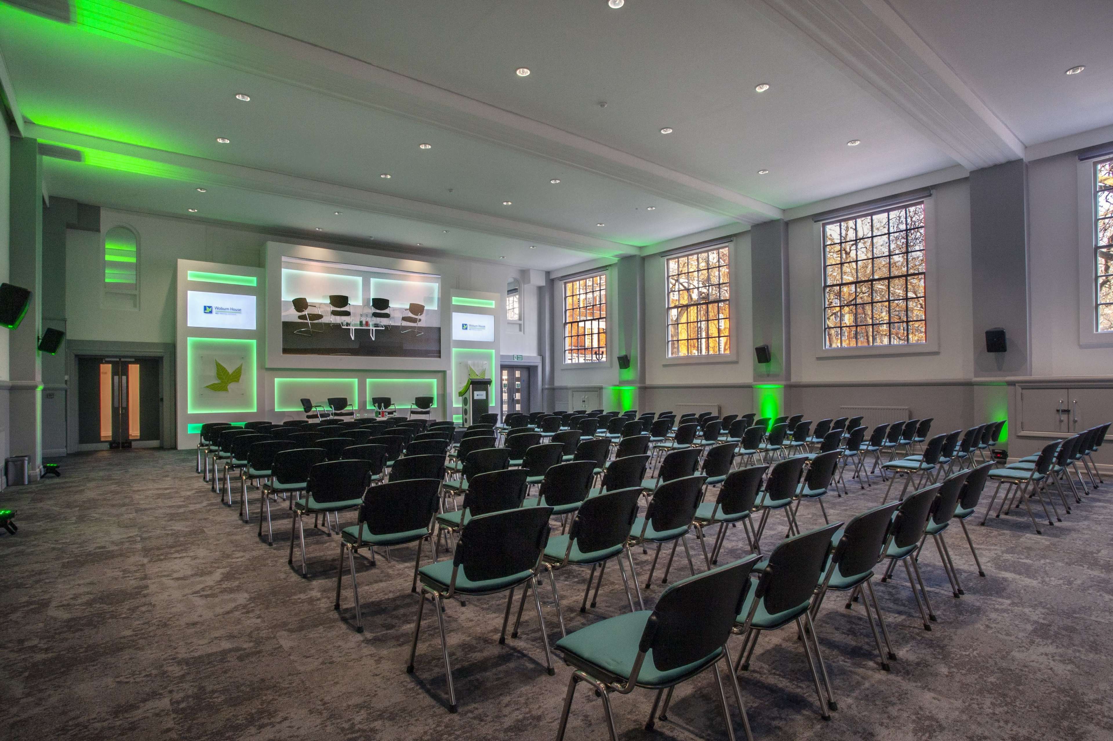 A conference room arranged with rows of black chairs facing a large presentation screen, illuminated by green accent lighting.
