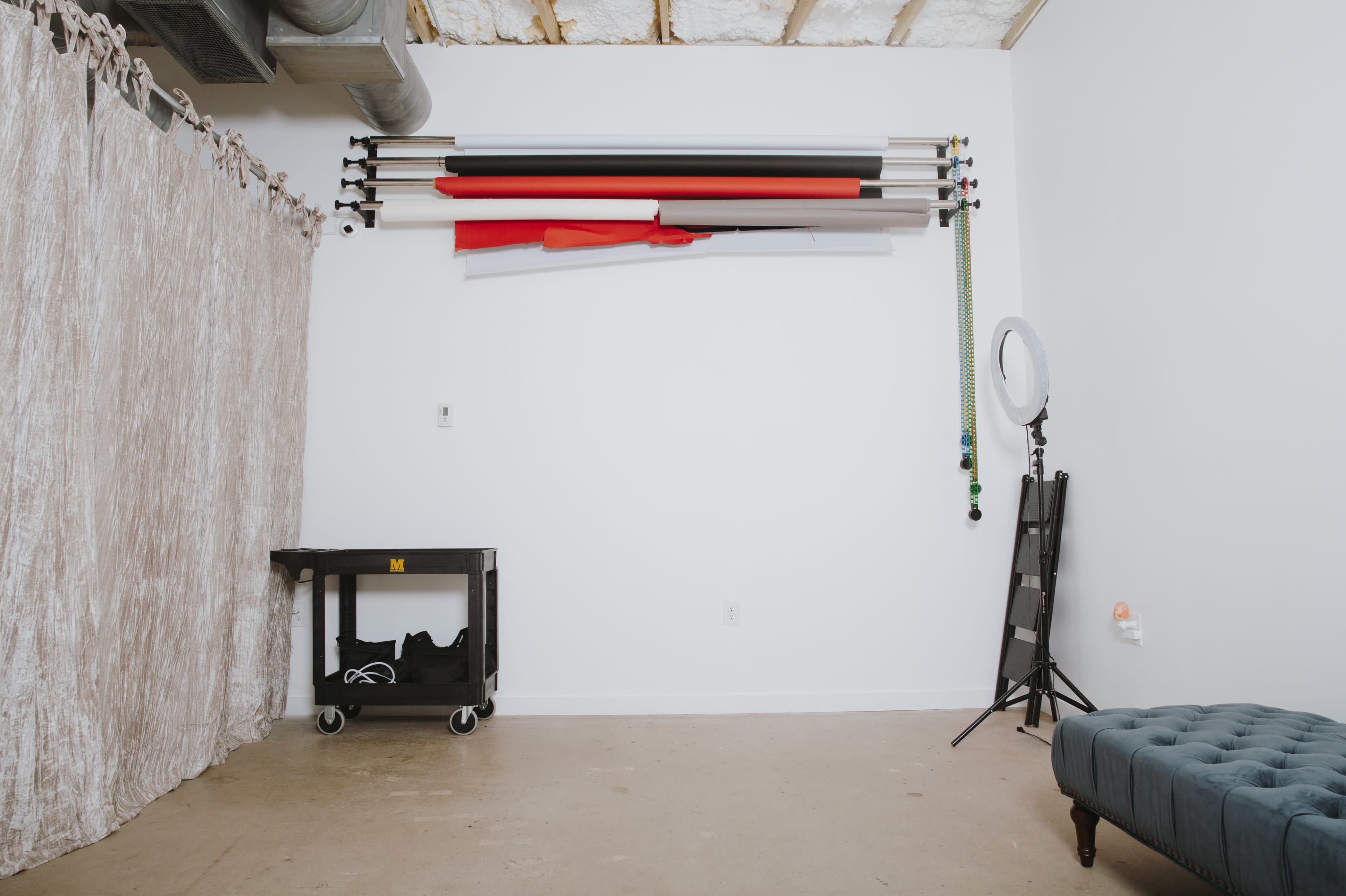 A minimalist studio interior with a wall of colored backdrop rolls, a small cart on wheels, a folding chair, and a ring light.