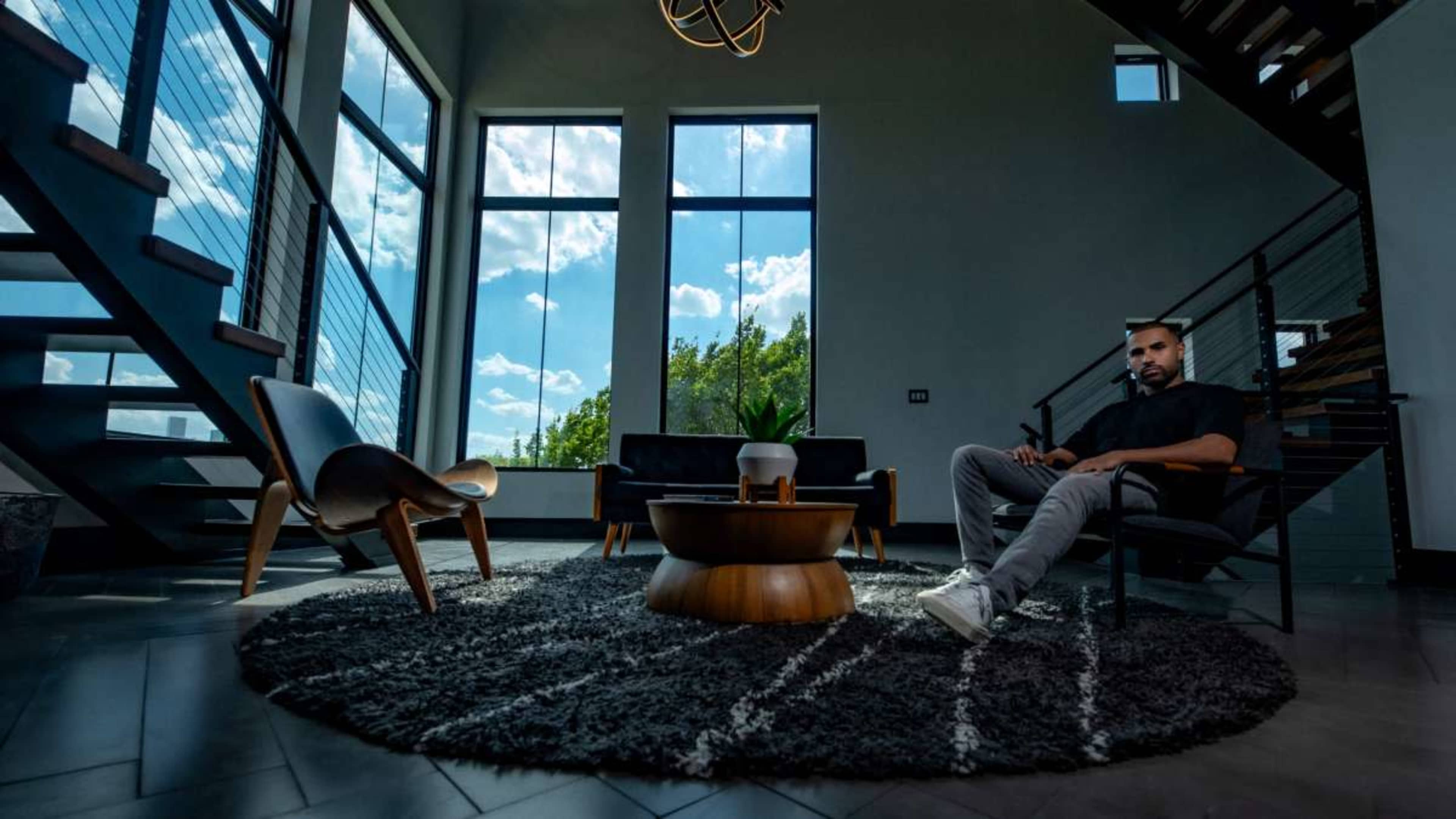 A man sits in a modern living room with large windows, displaying a view of the sky and greenery, alongside contemporary furniture.