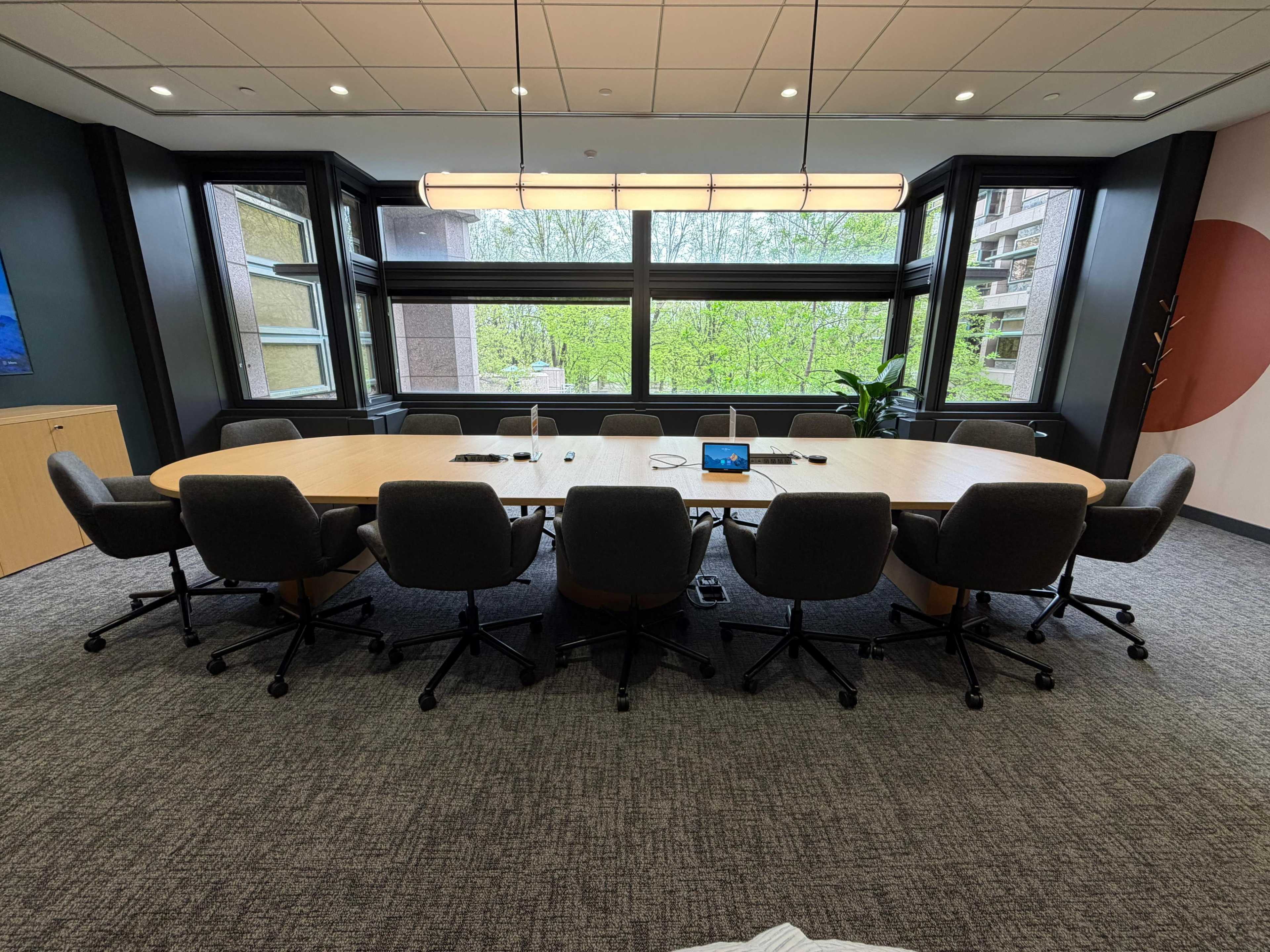 A large, oval conference table is surrounded by ergonomic chairs in a modern meeting room with large windows overlooking greenery.