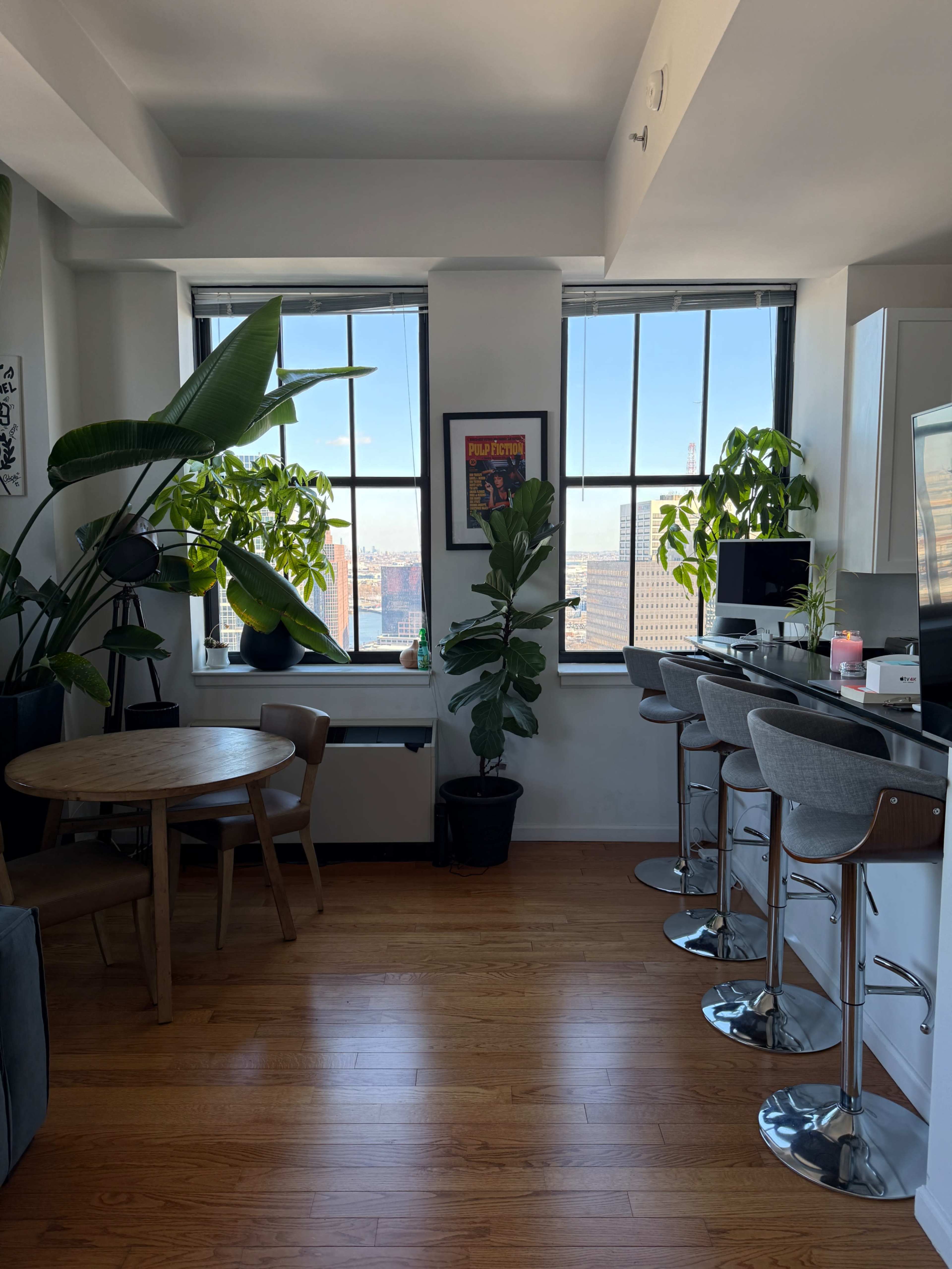 The image shows a modern apartment kitchen area with a round wooden dining table, high bar stools at a counter, and large windows featuring potted plants.