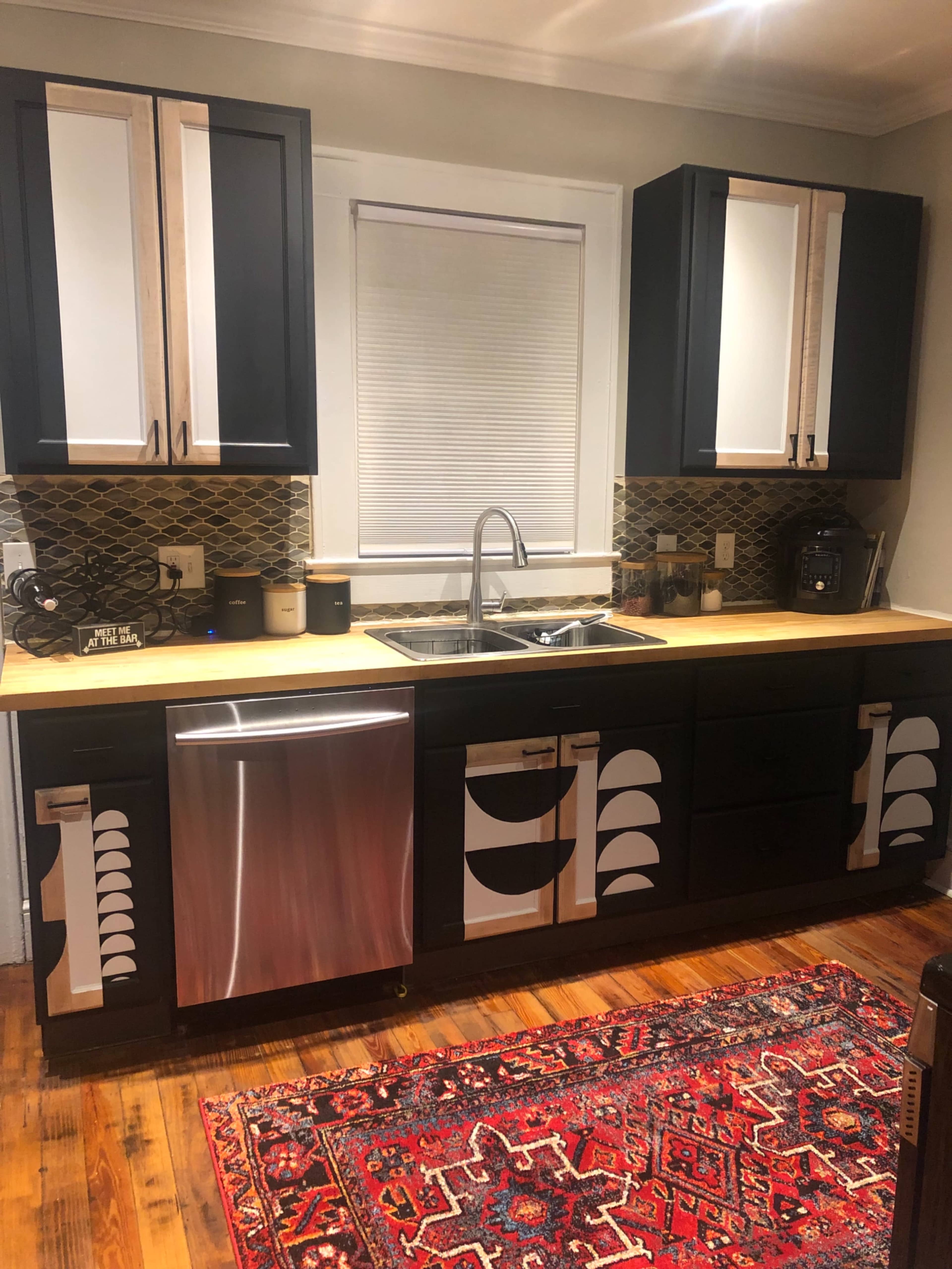 A kitchen with black and white cabinetry, a stainless steel dishwasher, a wooden countertop, and a decorative rug on the floor.