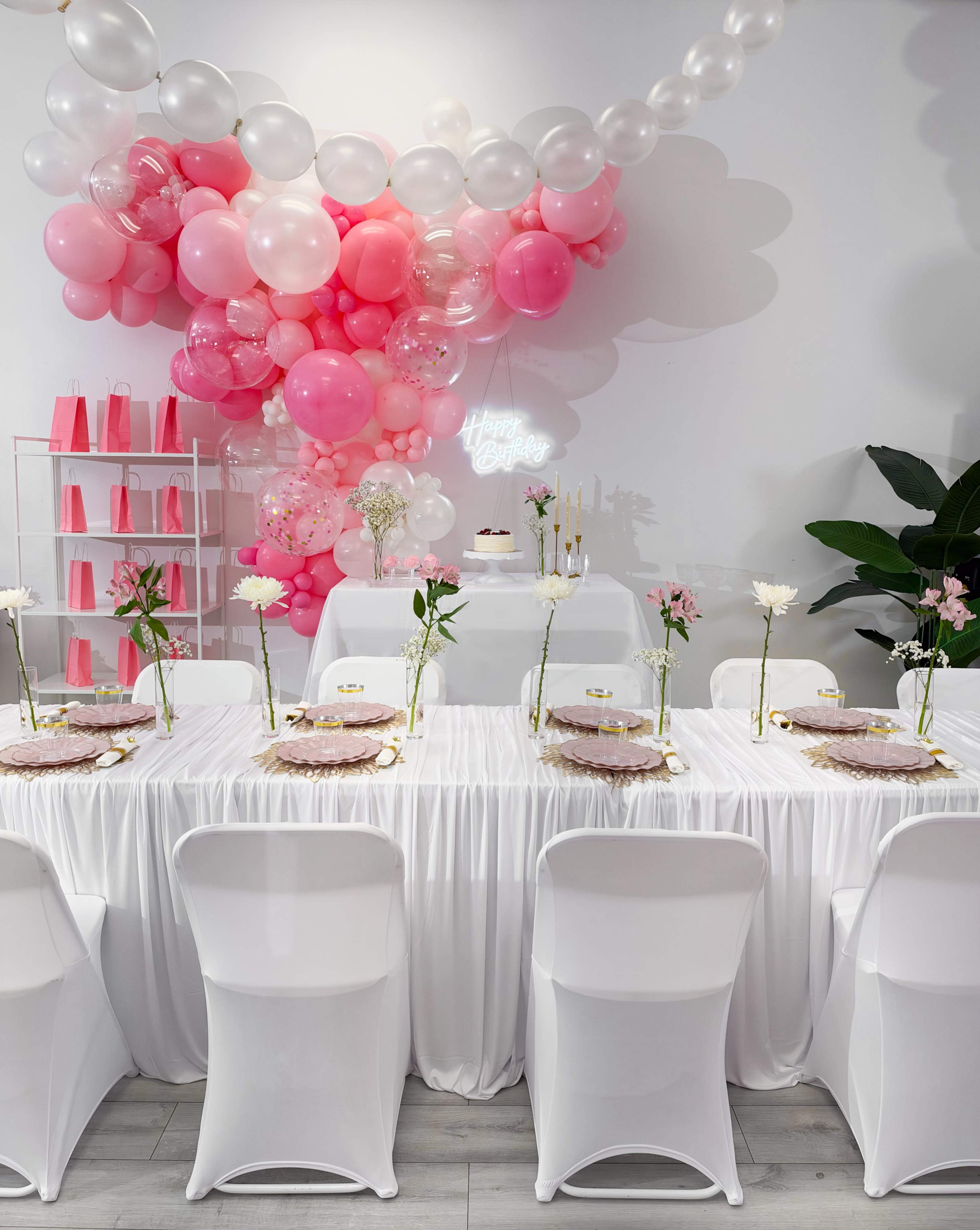 The image shows a decorated event space with a long dining table covered in a white tablecloth, adorned with floral centerpieces and surrounded by white chairs, alongside a backdrop of pink and white balloons.