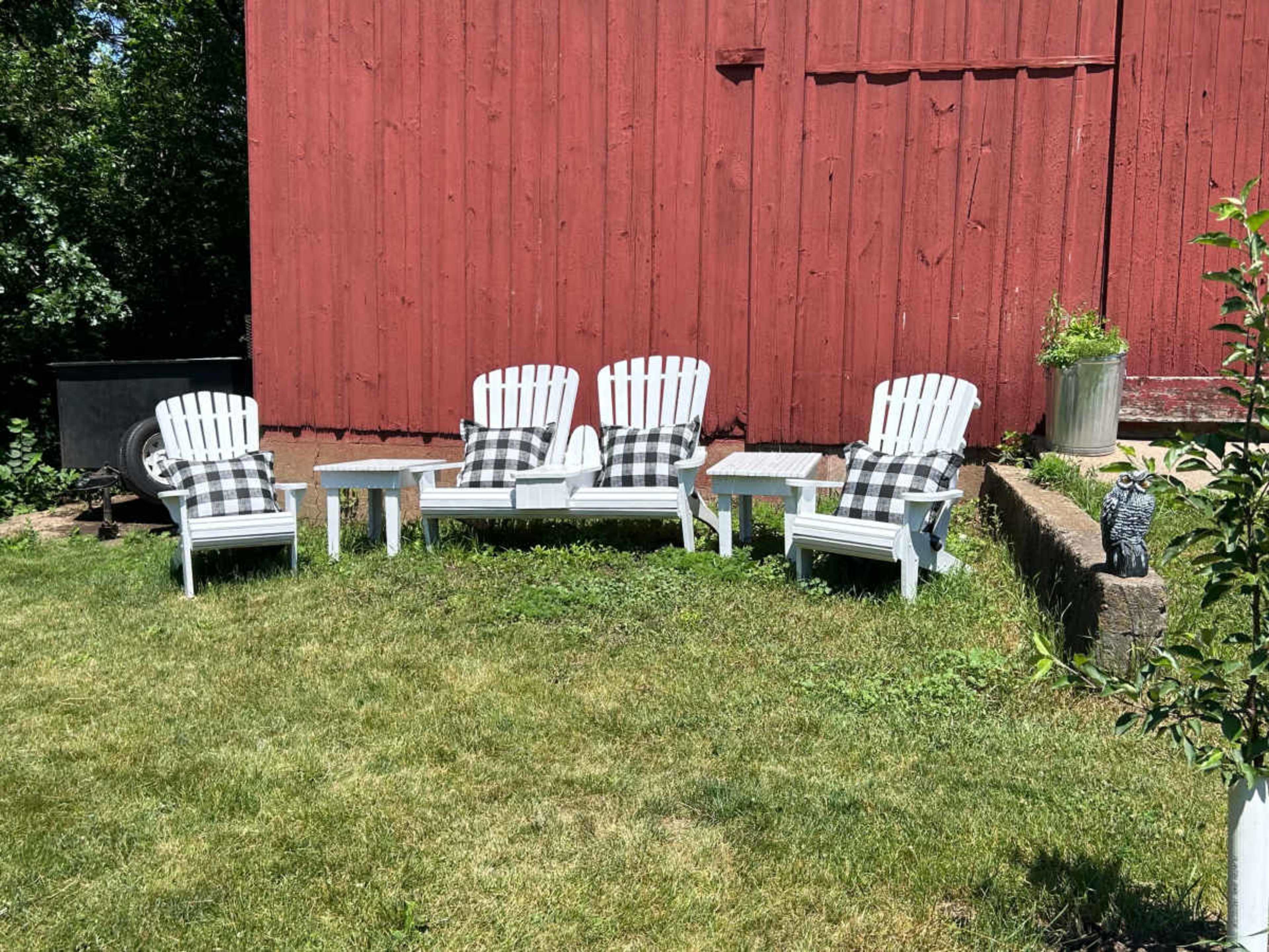 A set of four white Adirondack chairs with matching side tables is arranged on a grassy area in front of a red barn.